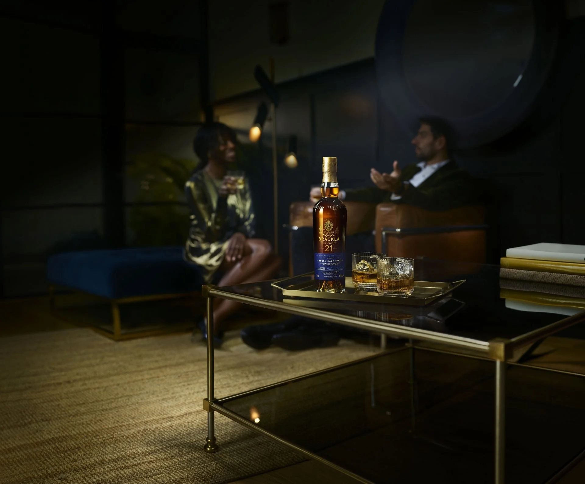 Two people sit and converse in a dimly lit room at night, with a bottle of Royal Brackla whiskey and two glasses of whiskey on a coffee table in front of them.