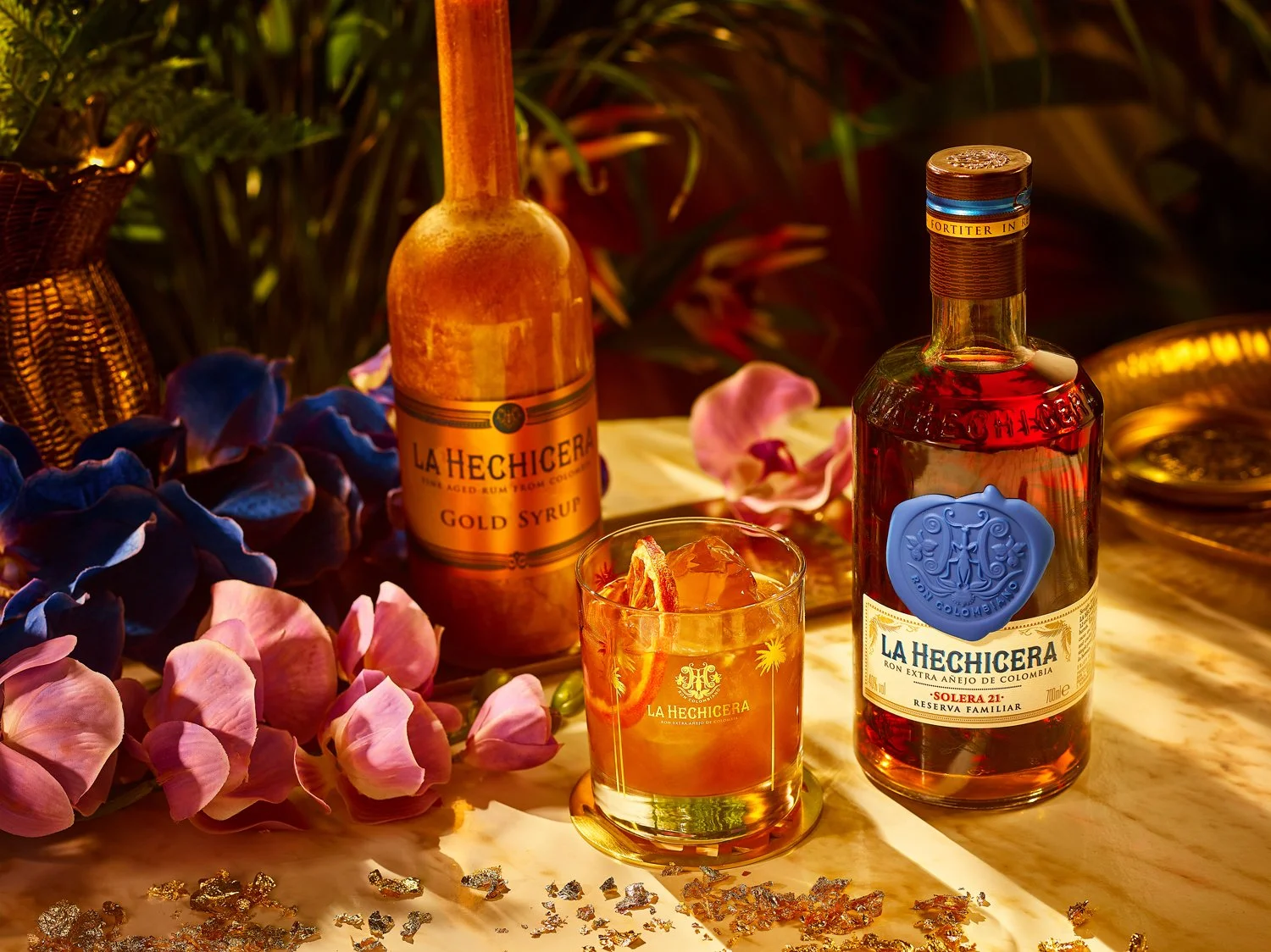 A bottle of La Hechicera rum, a glass of rum with ice and a slice of orange, surrounded by pink and blue flowers on a marble surface. There is also a bottle of La Hechicera Gold Syrup and a decorative gold plate in the background.