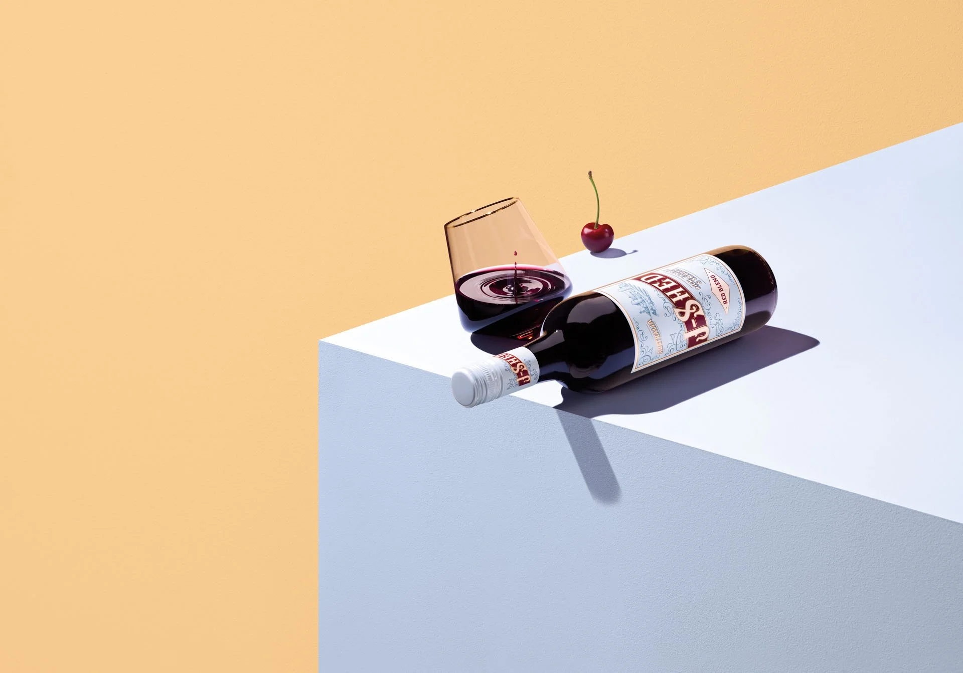 A bottle of red wine lying on its side on a white surface, with a glass of red wine and a cherry on top, against a two-tone pastel background.
