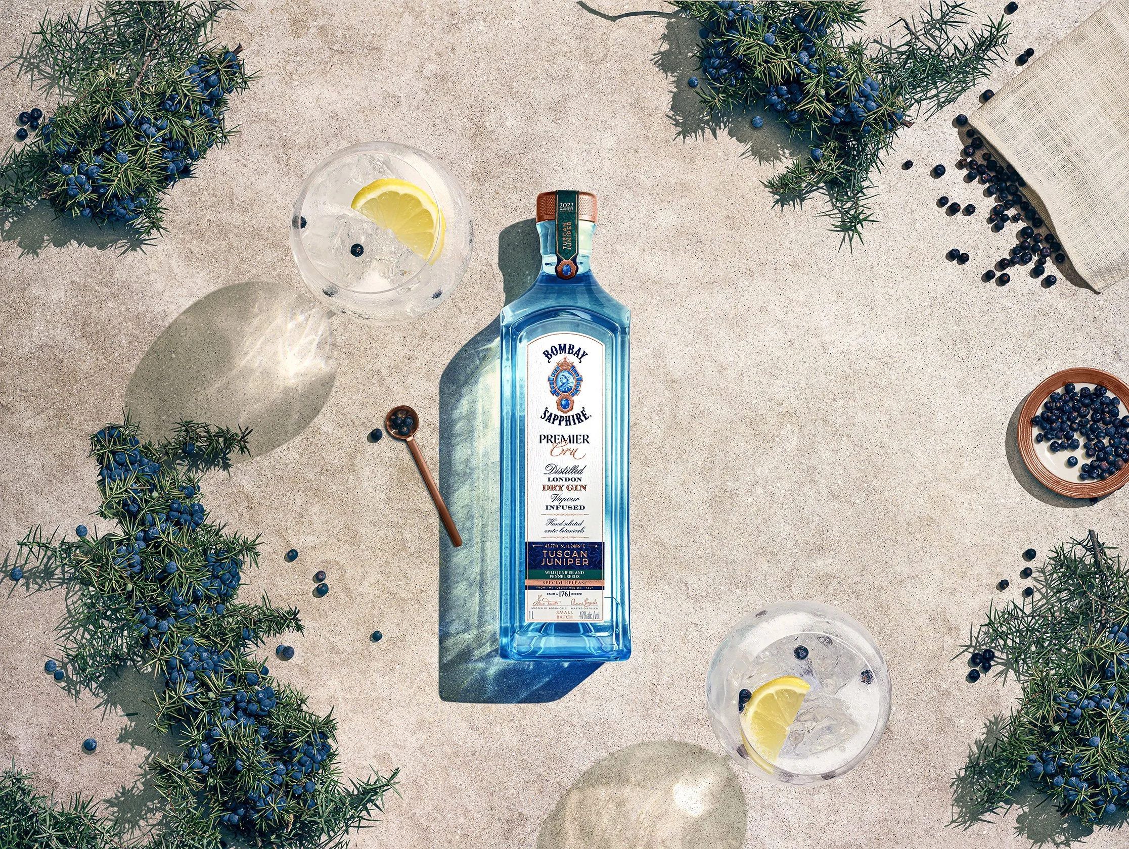 A bottle of Bombay Sapphire gin, two glasses with ice and lemon wedges, surrounded by green juniper branches with blue berries, on a sandy surface.