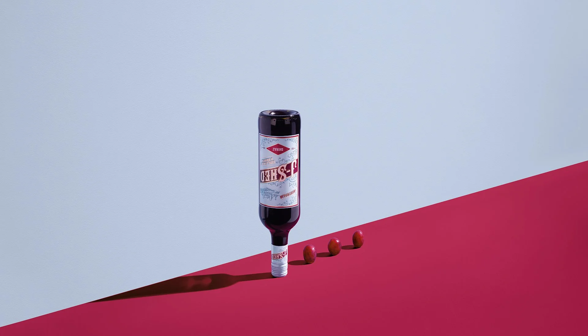 A row of red grapes, a vertically standing bottle of Shiraz wine, and a white wine cork on a red surface against a gray and red background.