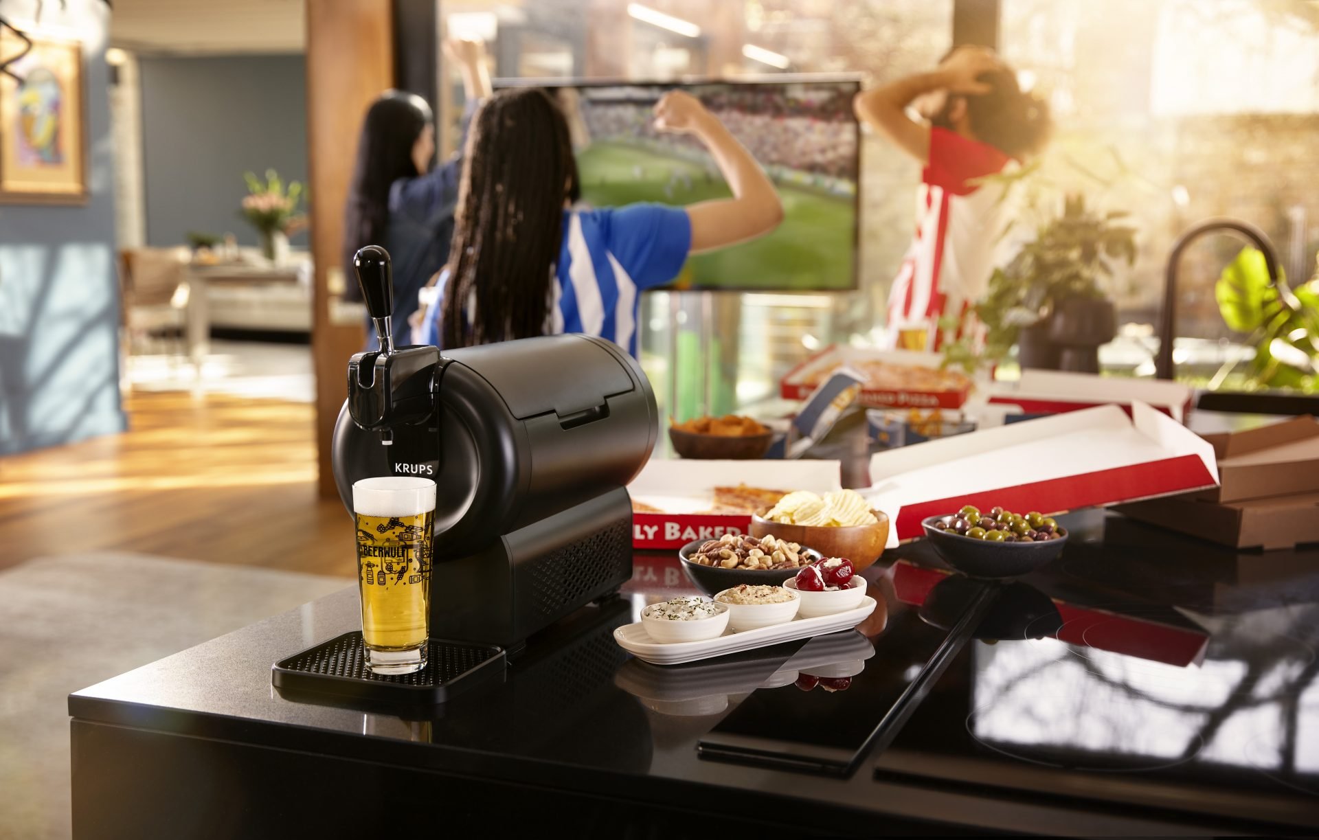 A kitchen counter with a Keurig coffee machine, a glass of beer, bowls of toppings including cherries, nuts, and whipped cream, pizza boxes, and snack foods, with a woman watching TV in the background.
