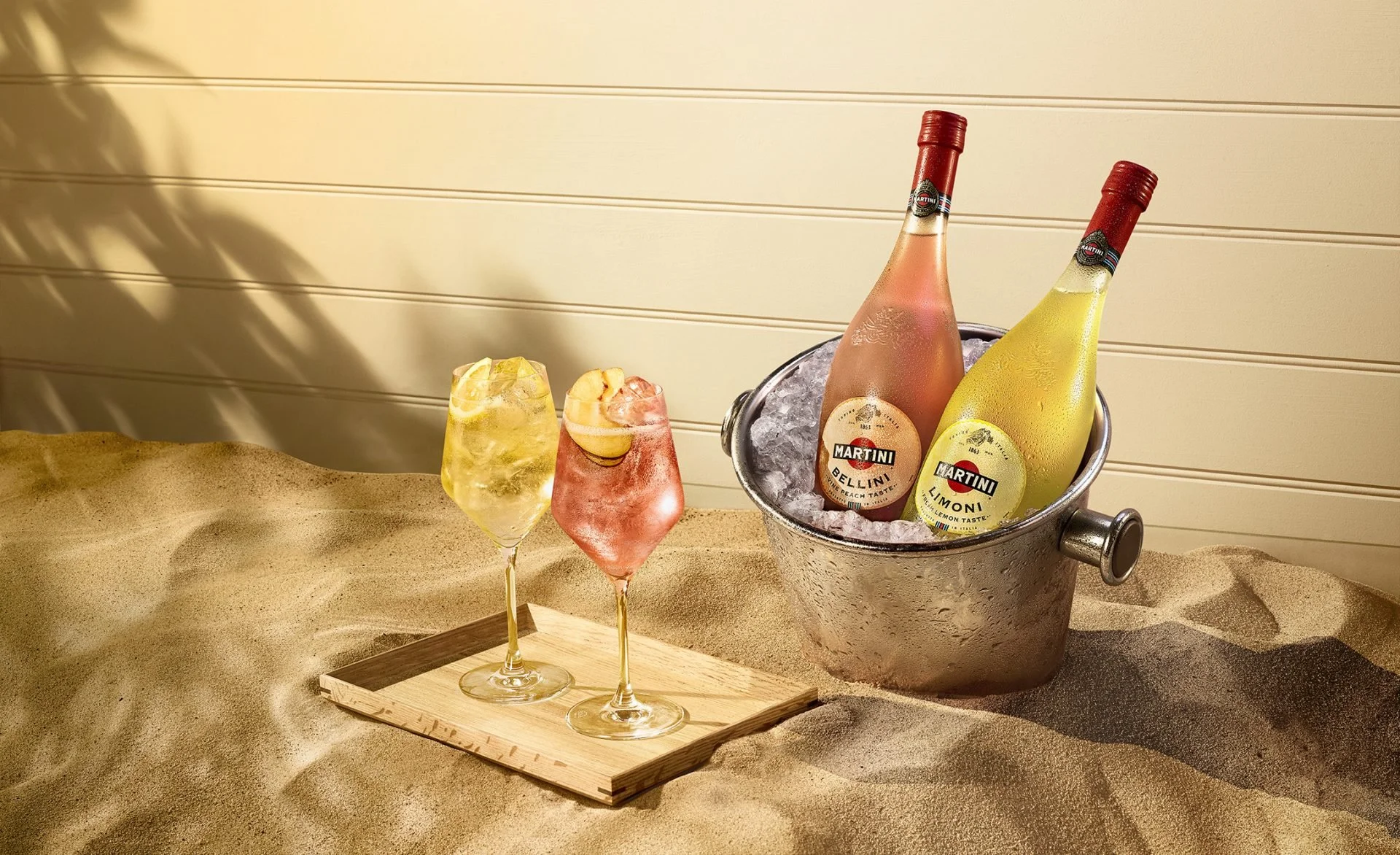 Two colorful cocktails with lemon slices on a wooden tray, and two bottles of Martini sparkling wine chilling in a silver ice bucket on a sandy surface.