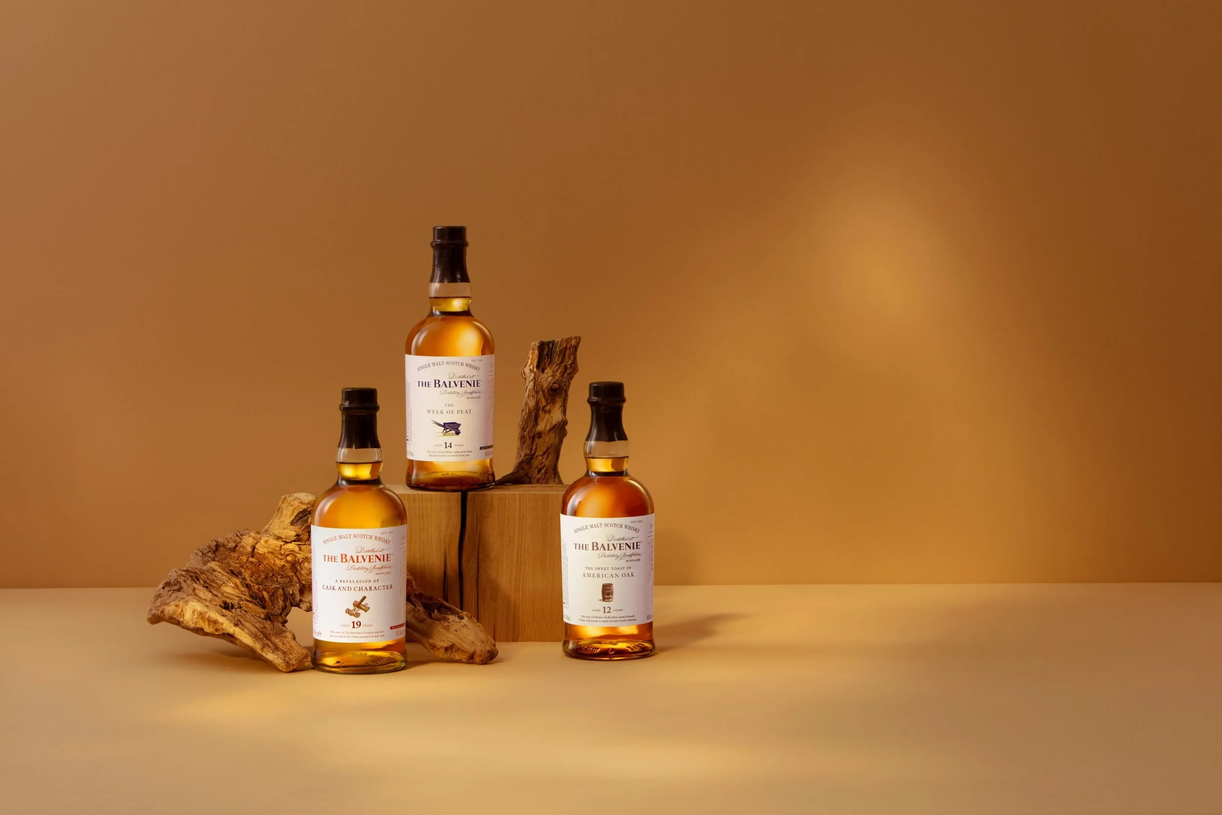 Four bottles of The Balvenie single malt Scotch whisky are arranged on a wooden surface with a piece of wood and a log. The background is a warm, solid color.
