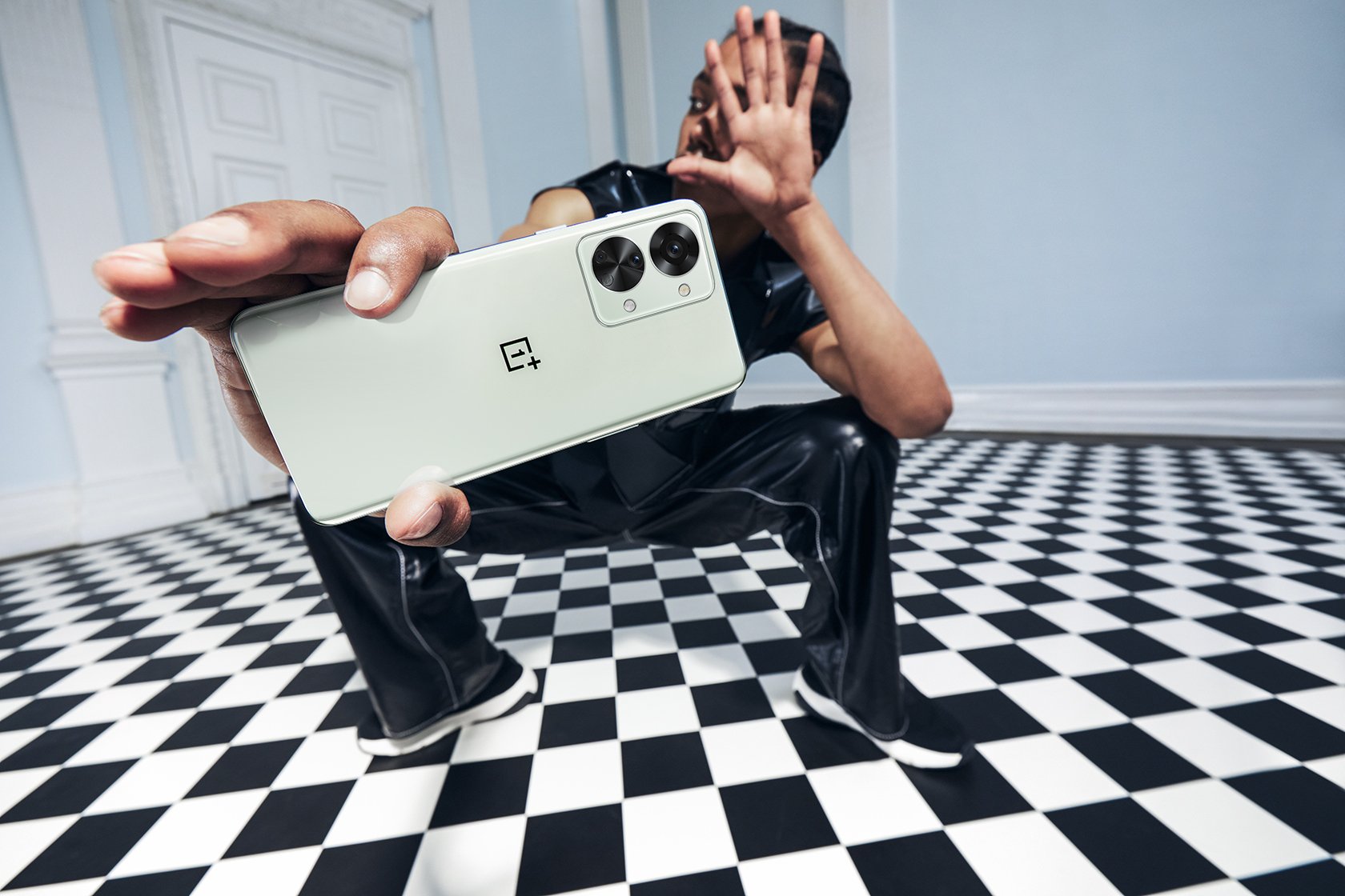 Person in black outfit sitting on black and white checkered floor, taking a selfie with a white OnePlus smartphone, with a hand extended toward the camera.