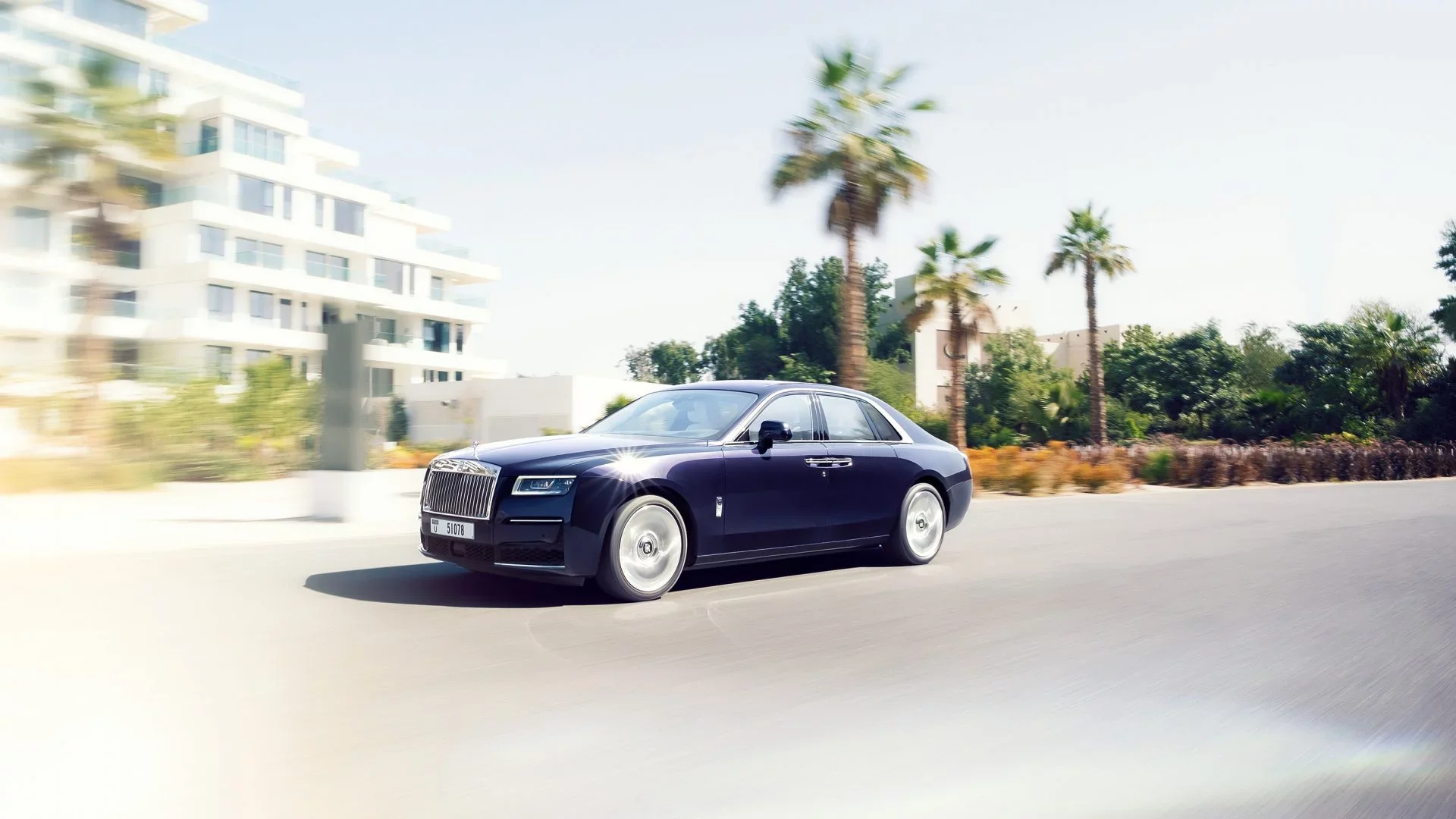 A sleek black luxury sedan driving on a road with palm trees and modern buildings in the background under a clear sky.
