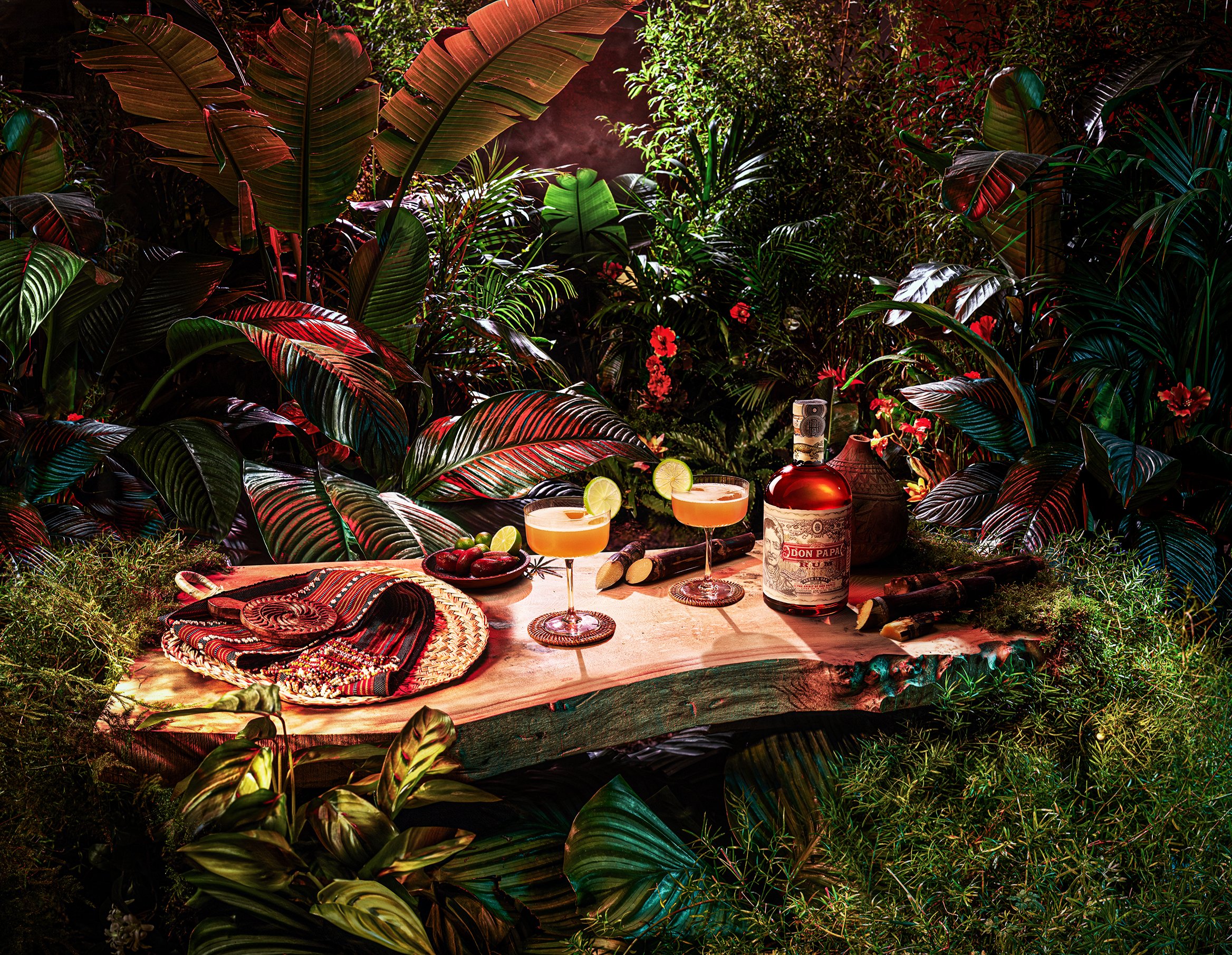 A lush, tropical jungle setting with large green and red leaves, and a table setup for drinks with two cocktails garnished with lime slices, a bottle of Don Papa rum, a bowl of strawberries, a small lemon, and various woven baskets and cloths on the 