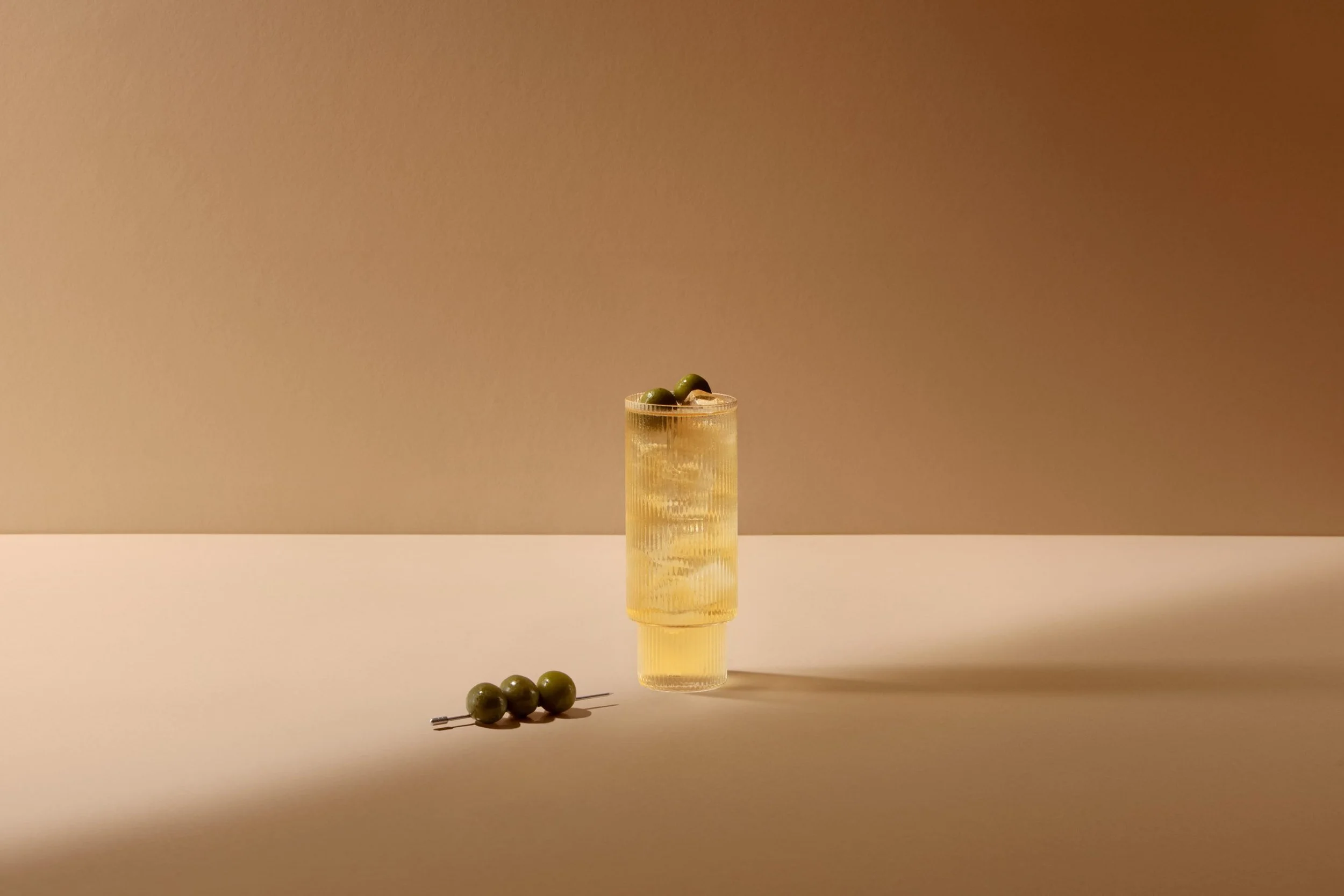 A tall glass of light yellow beverage garnished with green olives, with more green olives on a pin placed outside the glass and a small plate with additional olives on a minimalist surface against a beige background.