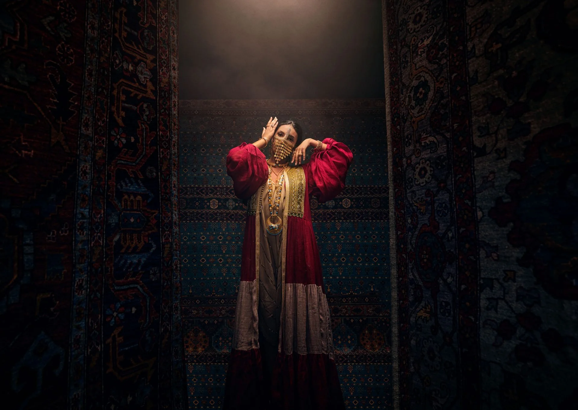 A woman dressed in traditional Indian attire, wearing a vibrant pink and gold outfit, ornate jewelry, and a decorative face mask, standing in a dimly lit room with intricate oriental rugs on the walls.