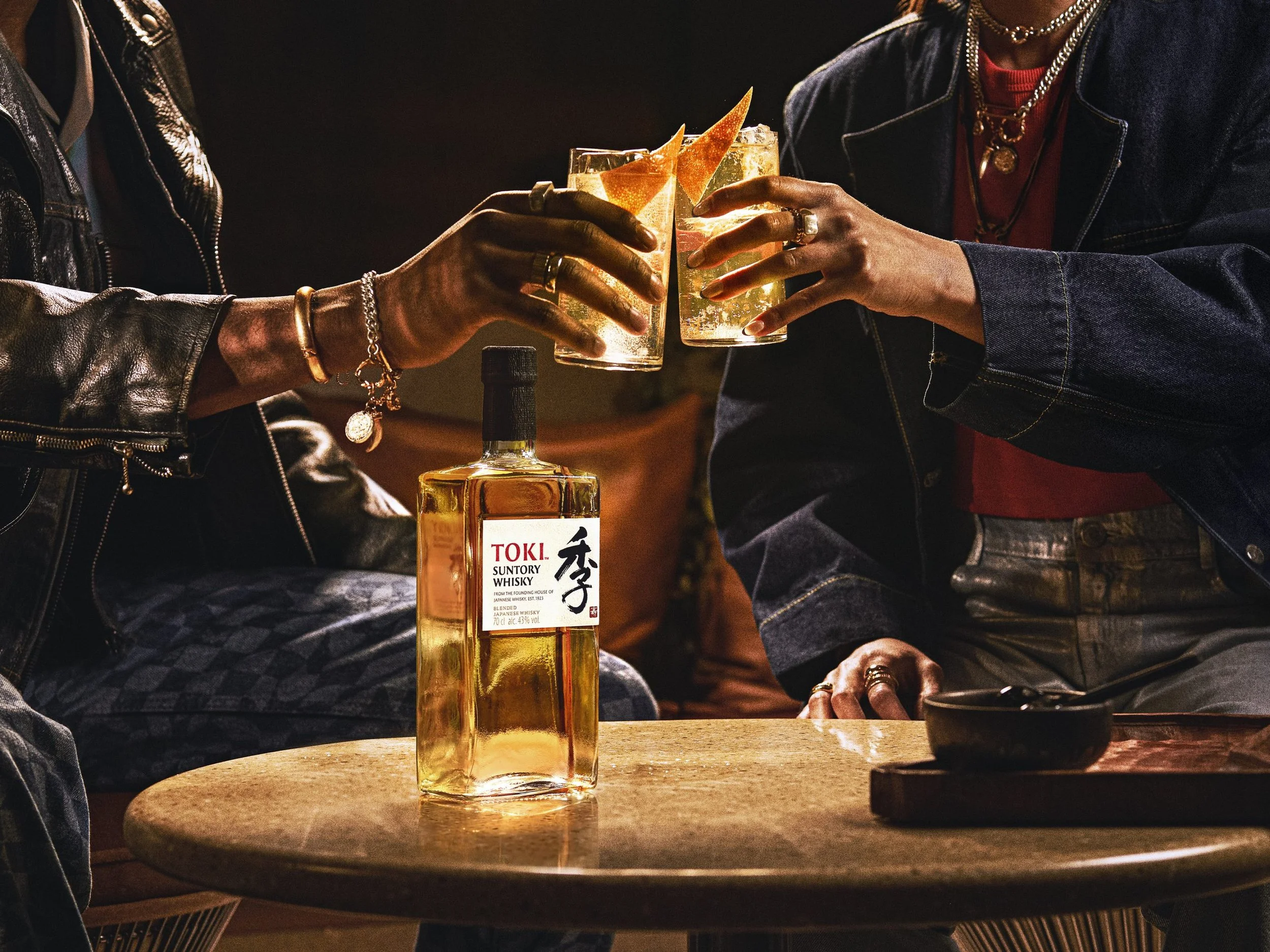 Two people clinking glasses of whiskey in a dimly lit setting. The glasses have garnishes, and there is a bottle of whiskey on the table.