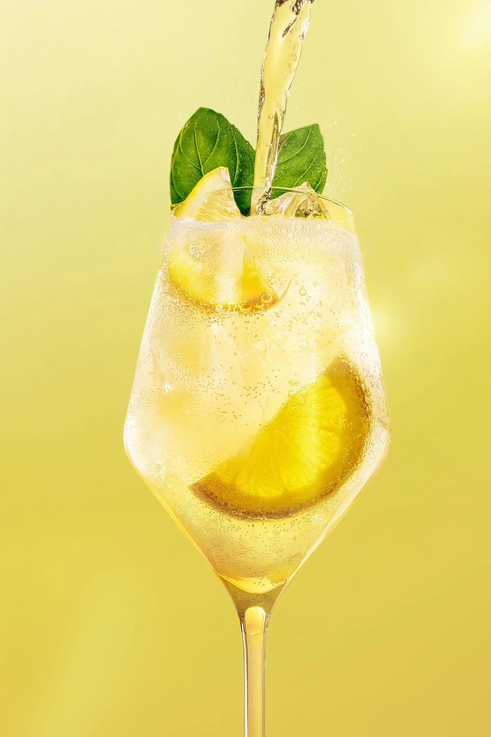 A glass of yellow beverage with lemon slices, ice cubes, and green basil leaves, being poured with liquid, set against a yellow background.