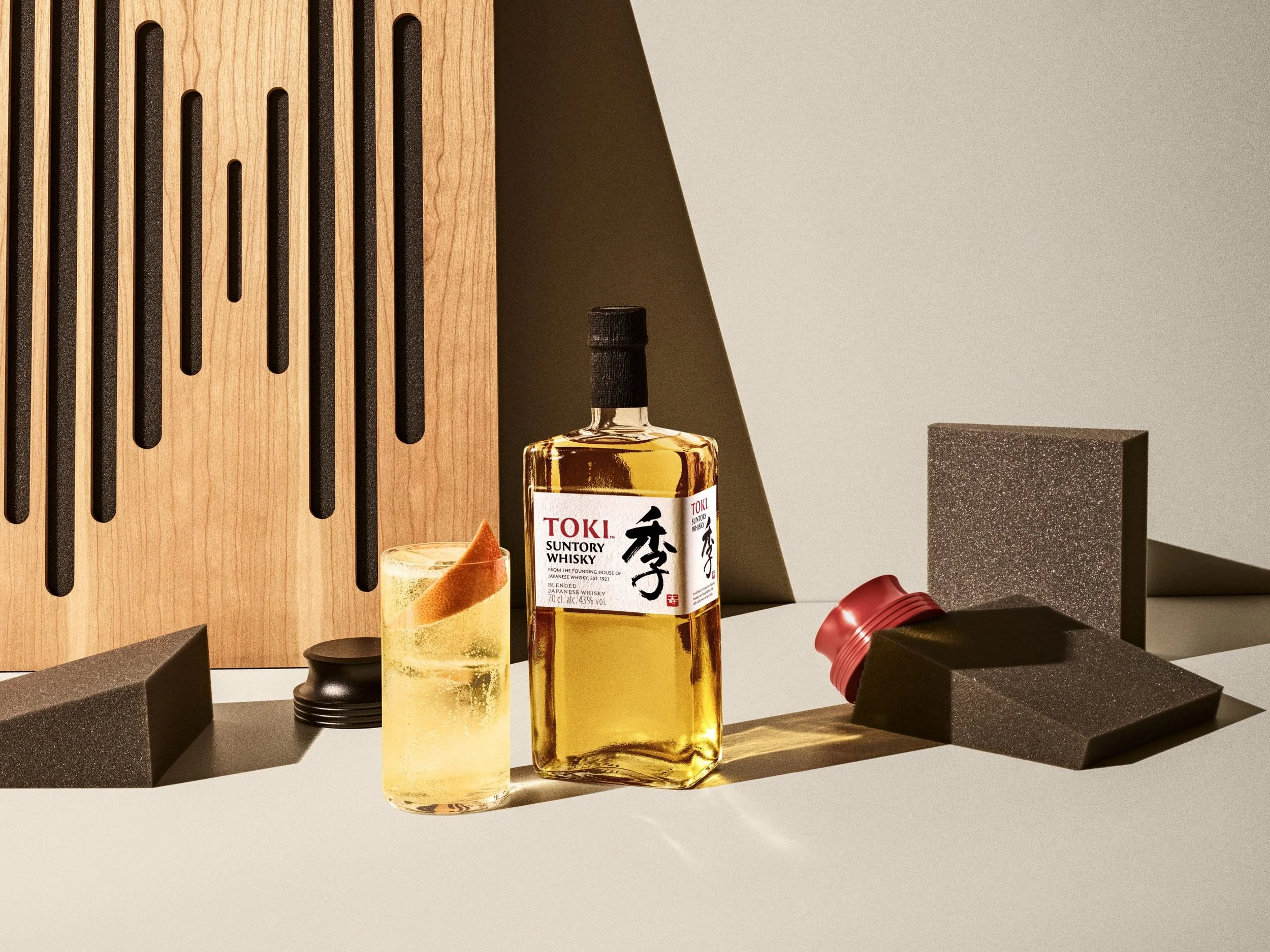 A bottle of Suntory Whisky, a glass with whisky and ice, and various black and brown foam blocks and other objects on a table, with a wooden slatted wall and brown background.