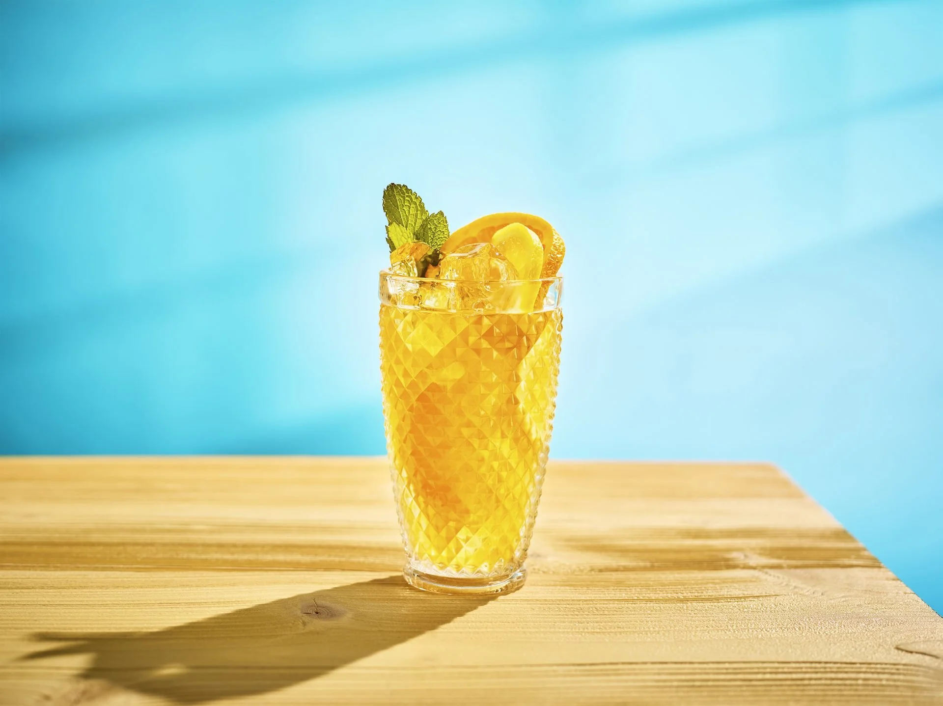 A tall glass filled with a yellow-orange beverage, garnished with lemon slices, mint leaves, and ice, placed on a wooden surface against a blue background.