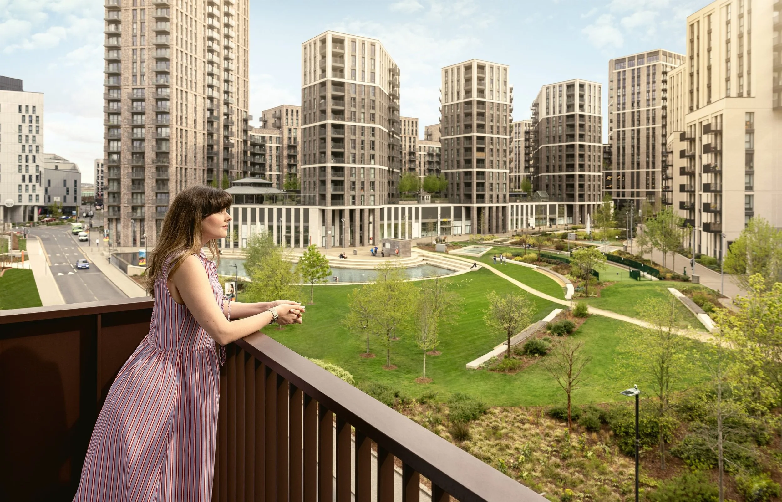 A woman with brown hair in a striped dress standing on a balcony overlooking a park and modern high-rise apartment buildings.