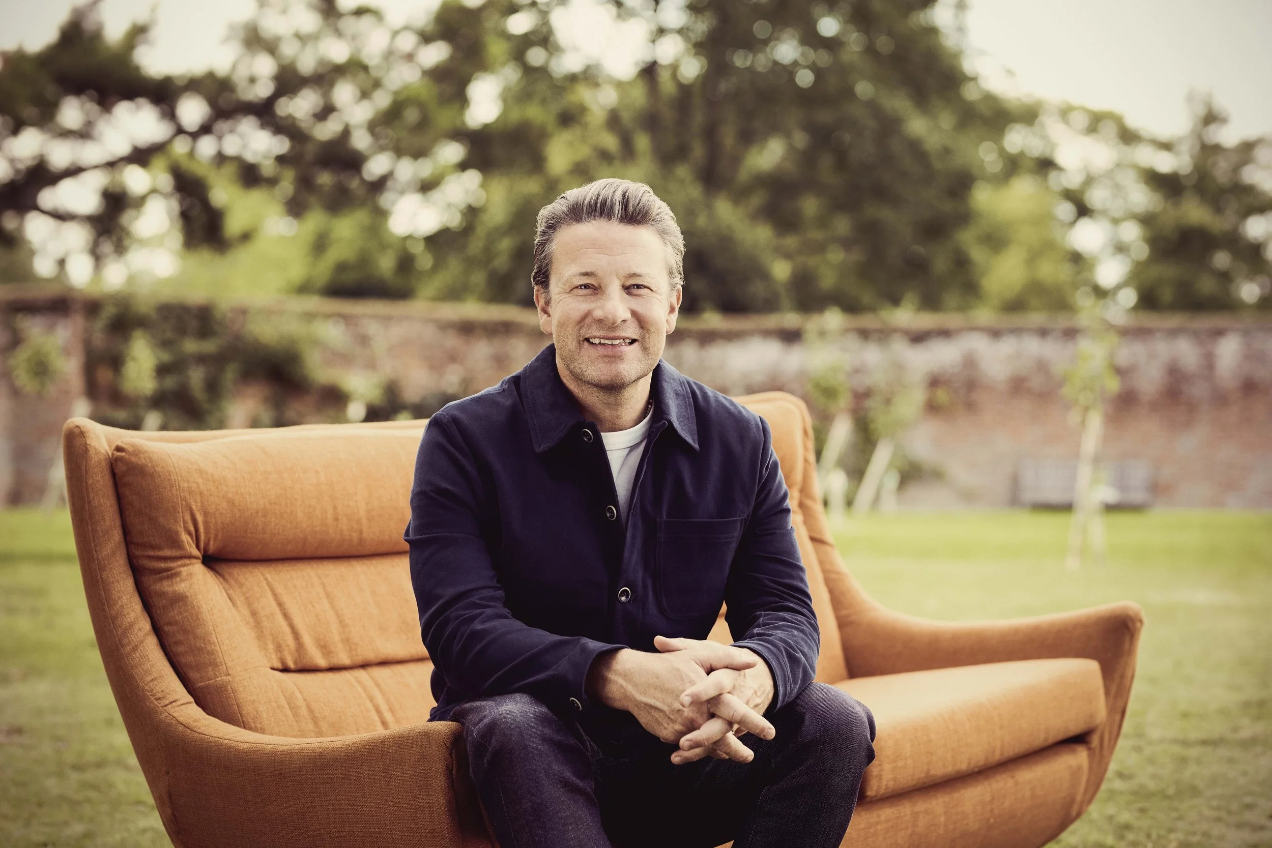 A smiling man with salt-and-pepper hair sitting on an orange outdoor sofa in a garden, with trees and a brick wall in the background.