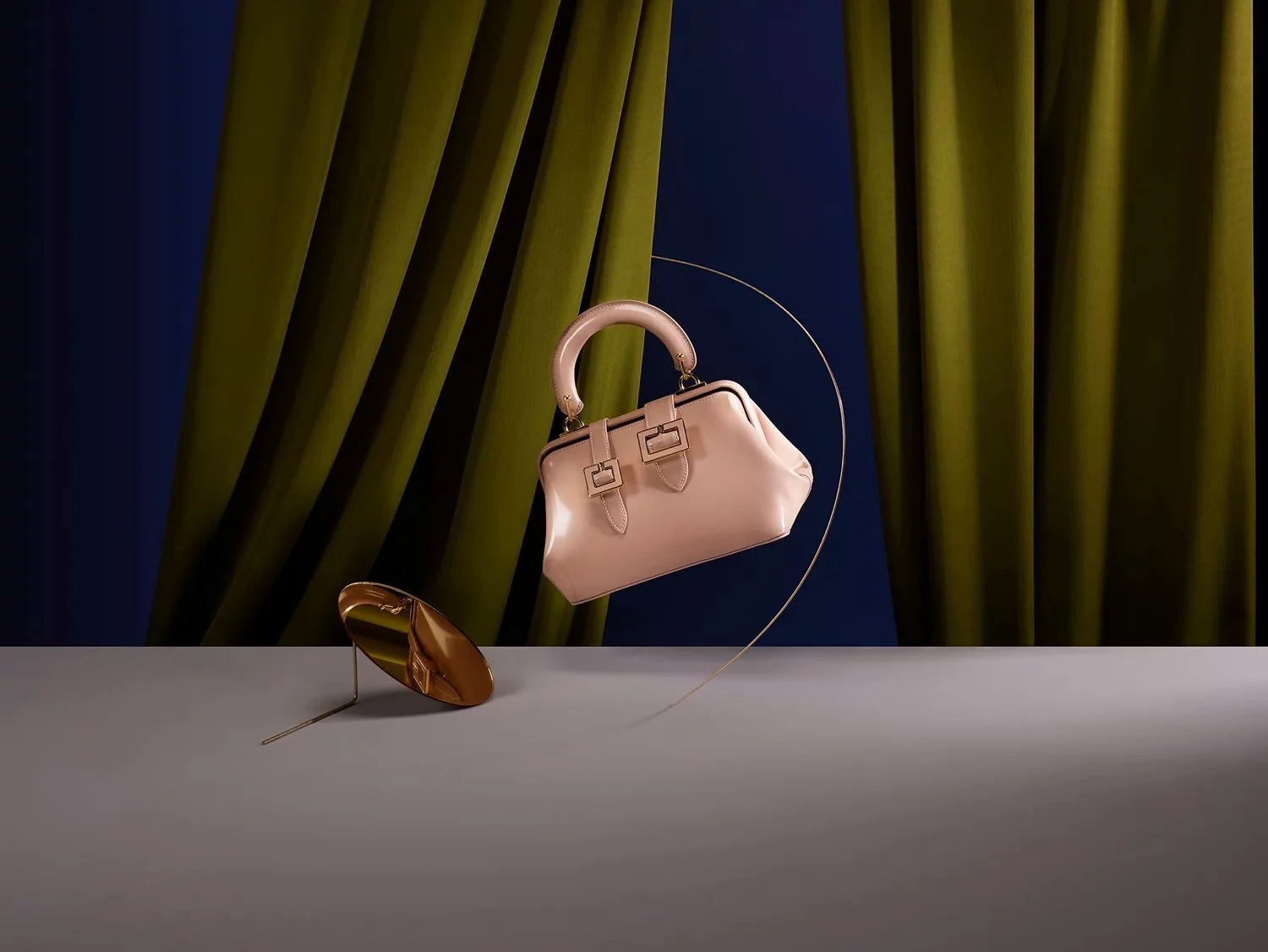 A light pink handbag with decorative buckles hangs from a thin gold curved stand, with a gold clutch purse resting on its side on a gray surface. The background features dark blue and greenish-yellow curtains.