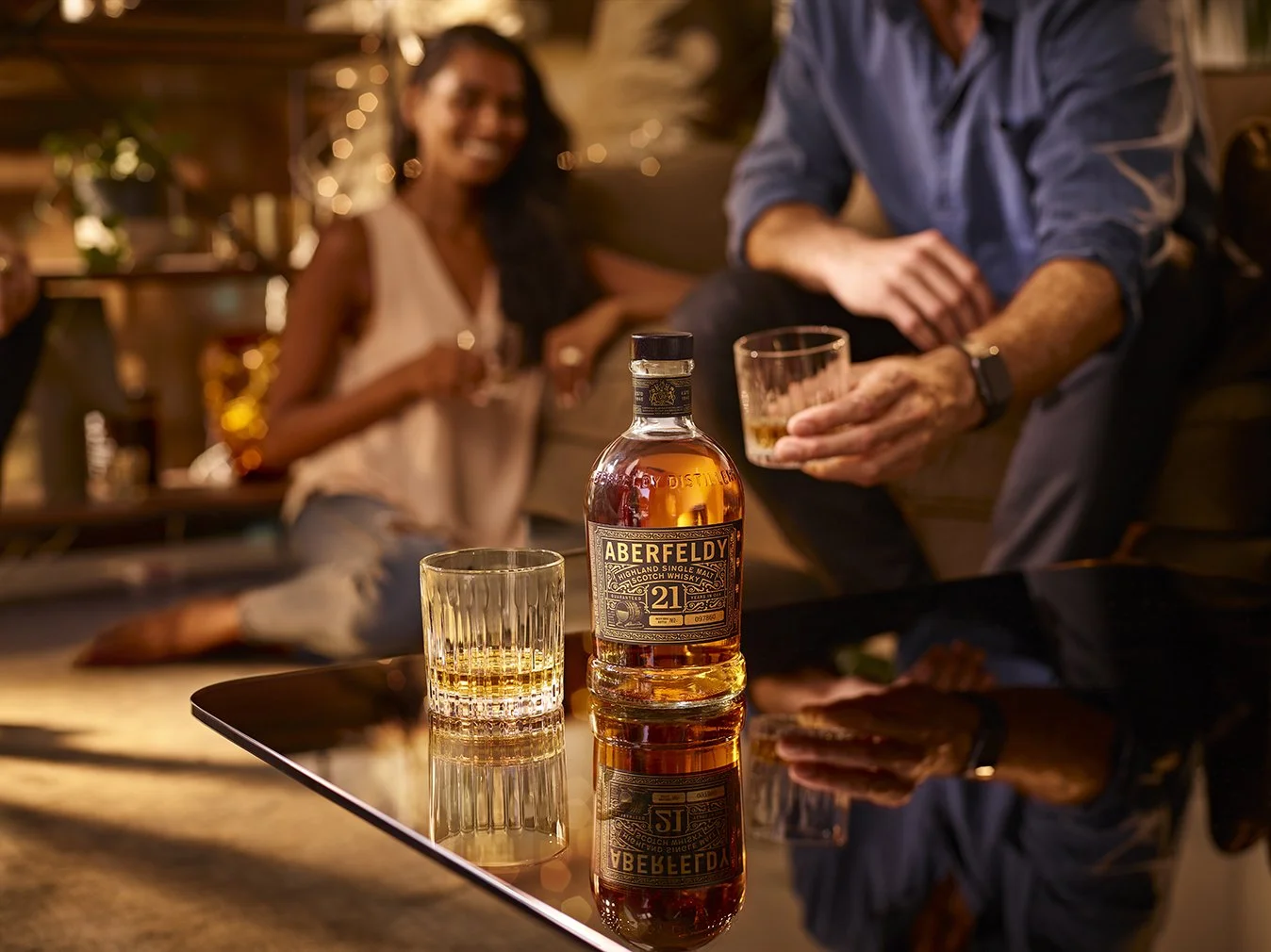 A bottle of Aberfeldy single malt whiskey and two glasses of whiskey on a reflective black surface in a cozy, dimly lit bar. In the background, a man and woman sit together, smiling and holding drinks, with blurred background lighting.