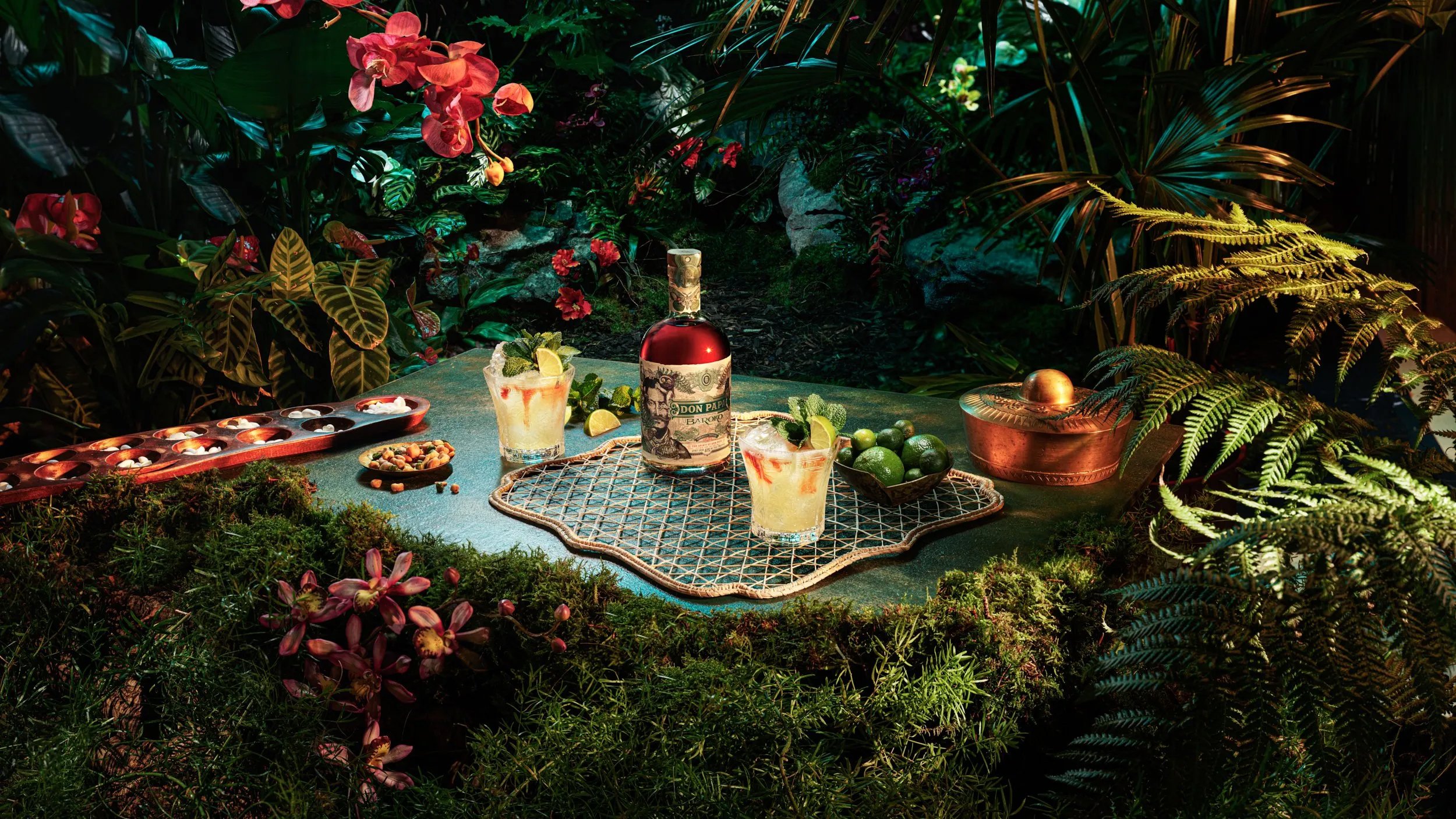 A tropical outdoor setting with a table decorated with drinks, lime, and small desserts, surrounded by lush green plants and flowers, illuminated with vibrant lighting.