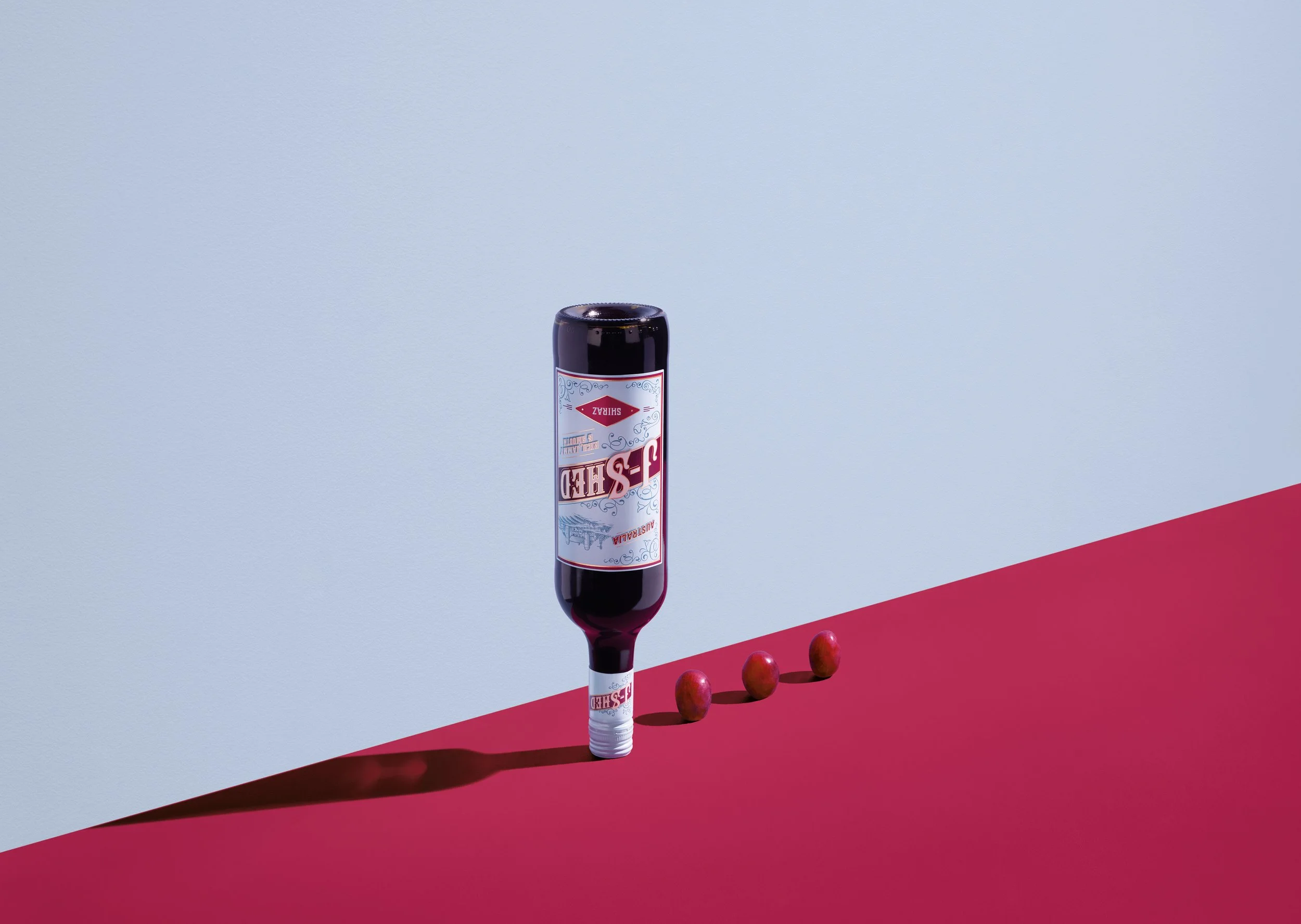 A bottle of Shiraz wine with a wine glass placed upside down on a pink and blue surface, with three cherries arranged in a row beside the glass.