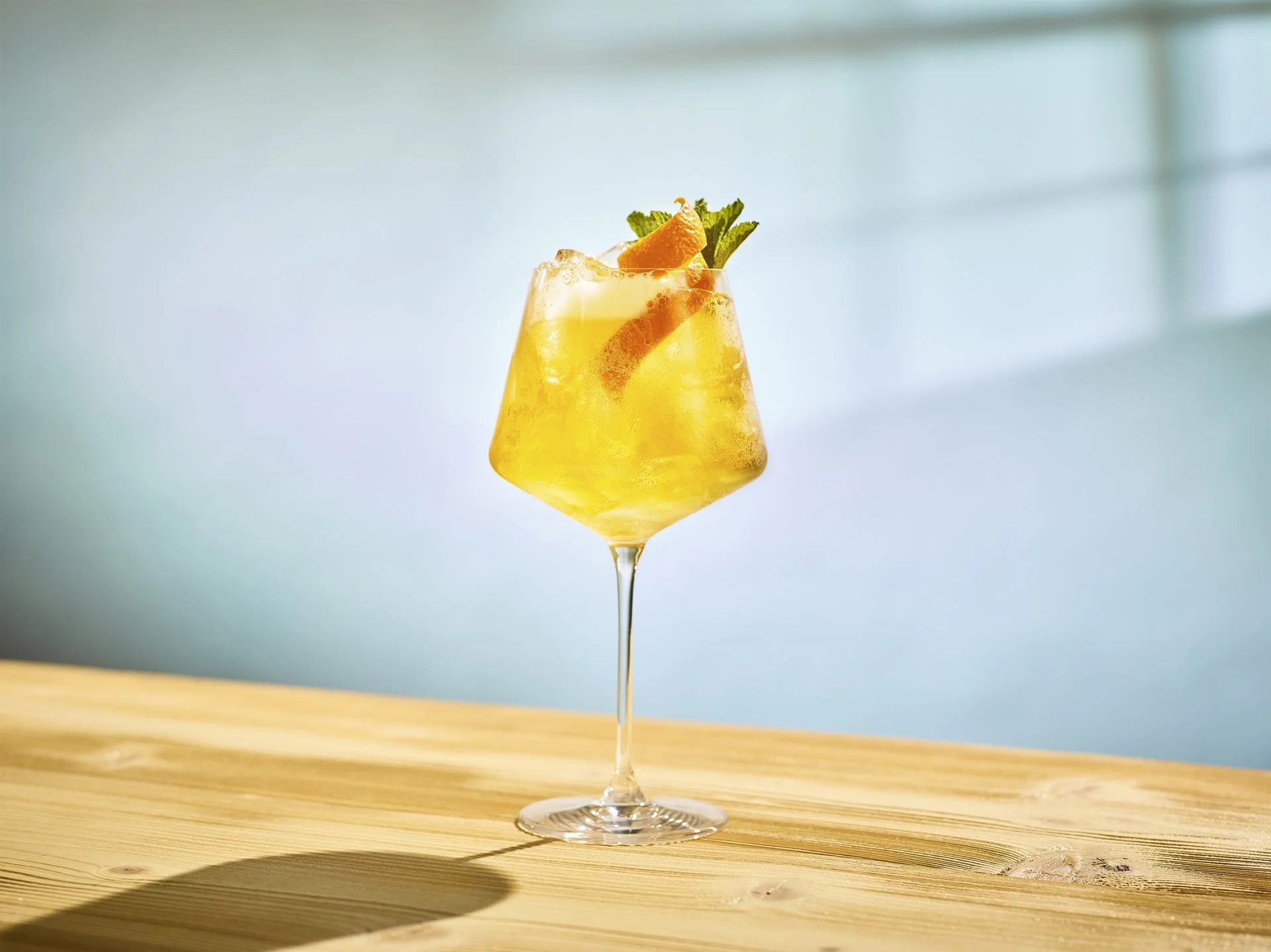 A glass of yellow cocktail garnished with orange slices, mint leaves, and a lemon peel on a wooden surface.