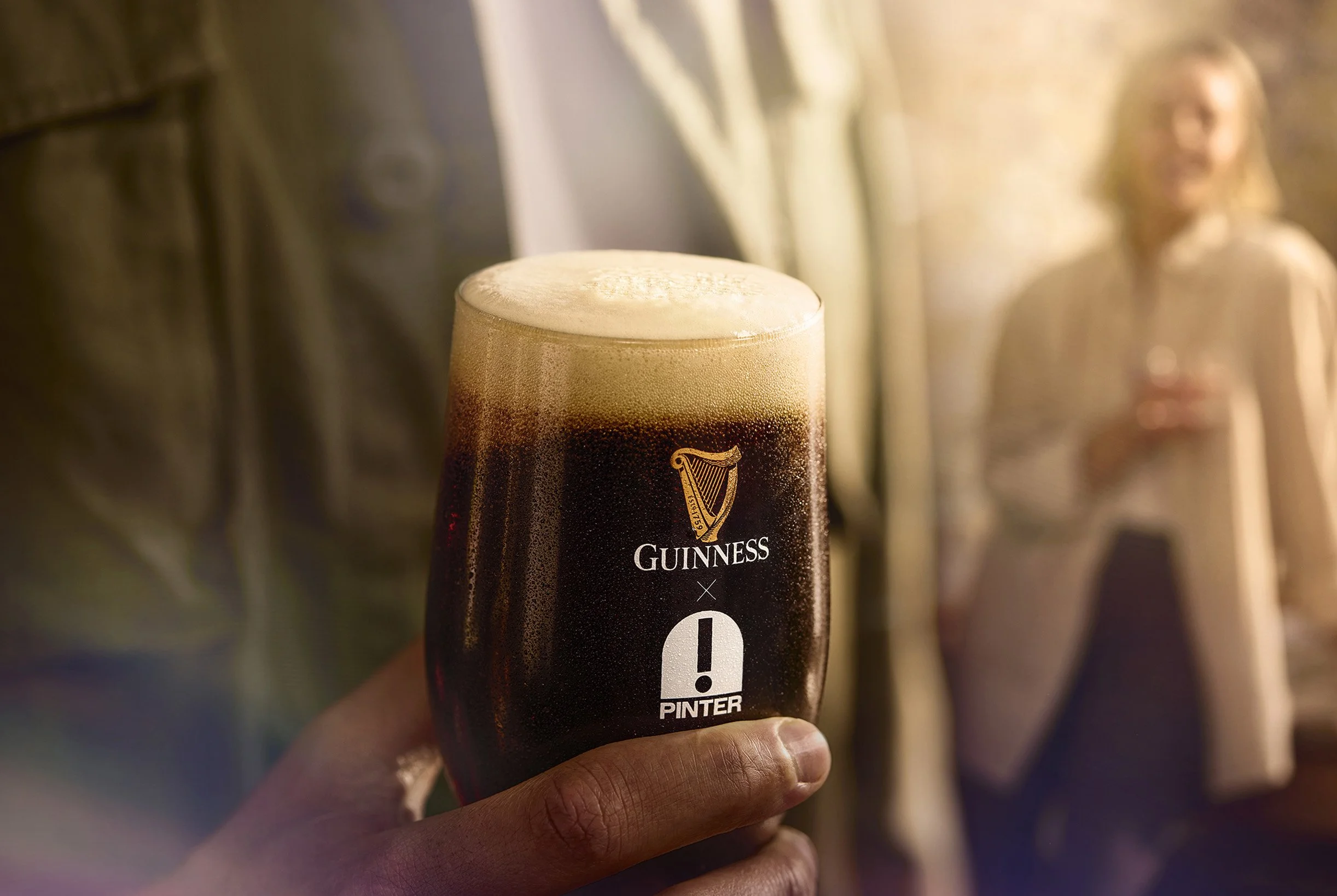 Close-up of a hand holding a pint glass filled with dark Guinness beer with a frothy head. In the background, a woman with curly hair and a striped shirt is seen smiling and talking to another person.
