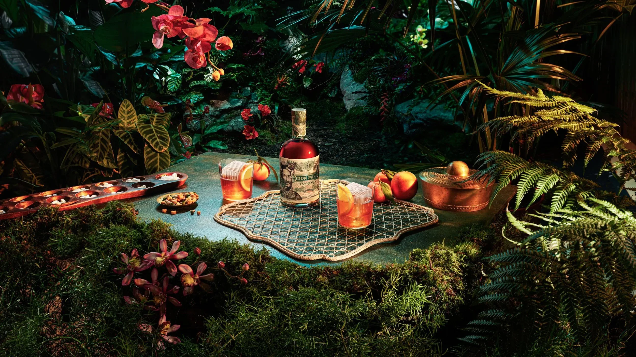 Tropical outdoor table setting with a bottle of Don Papa rum, two glasses of cocktail, apples, and snacks, surrounded by lush green plants and flowers.