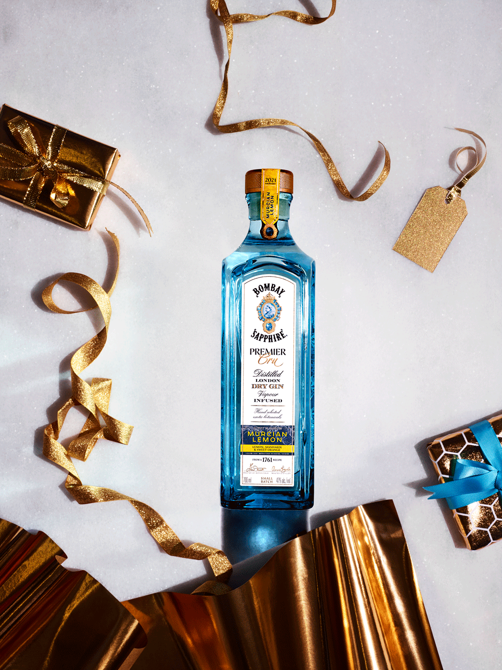 A bottle of Bombay Sapphire gin surrounded by gold and blue wrapped gifts, gold and glittery ribbons, and a gold tag on a white background with festive decorations.