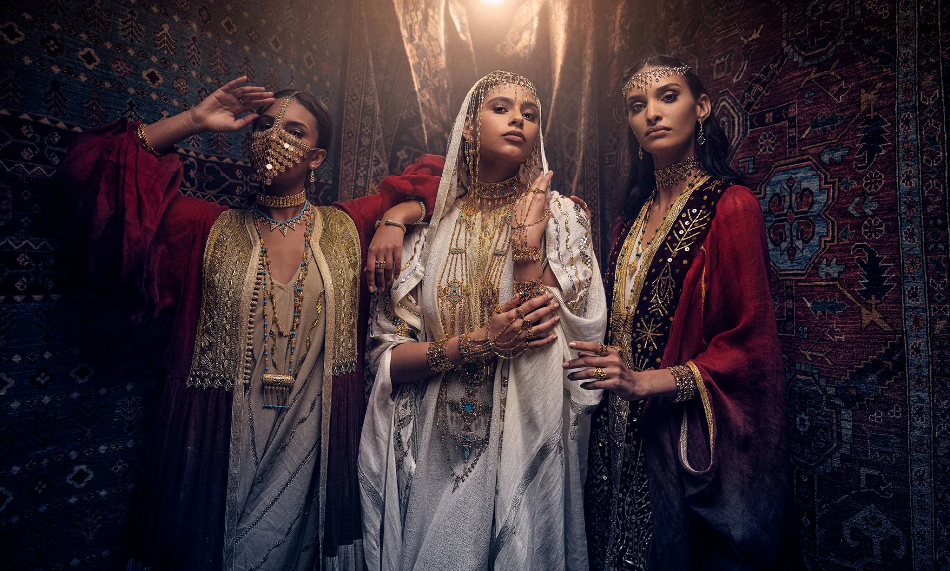 Four women wearing traditional and ornate jewelry and clothing, posing in front of a patterned carpet backdrop.