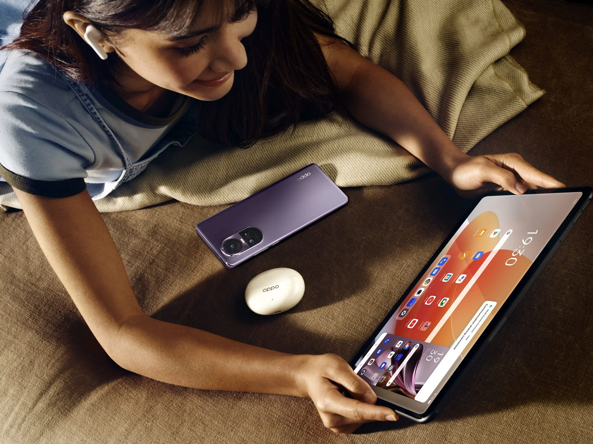 A woman lying on a beige couch using a tablet, with a purple smartphone and a white wireless earbud case nearby. She is wearing a gray shirt and has dark hair.