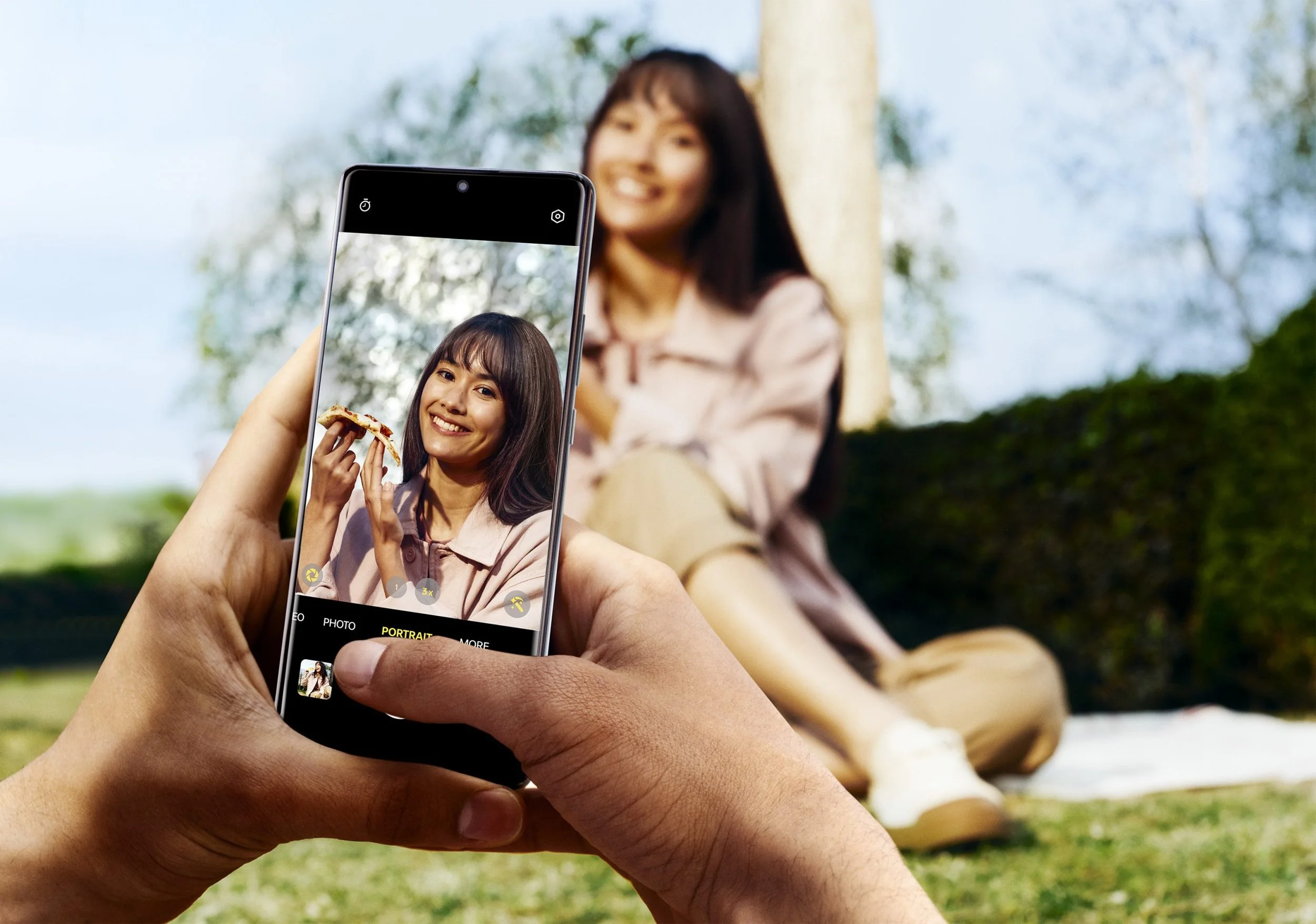 Person taking a photo of a woman sitting outdoors with a smartphone, woman smiling and holding a slice of pizza, blurred background with trees.