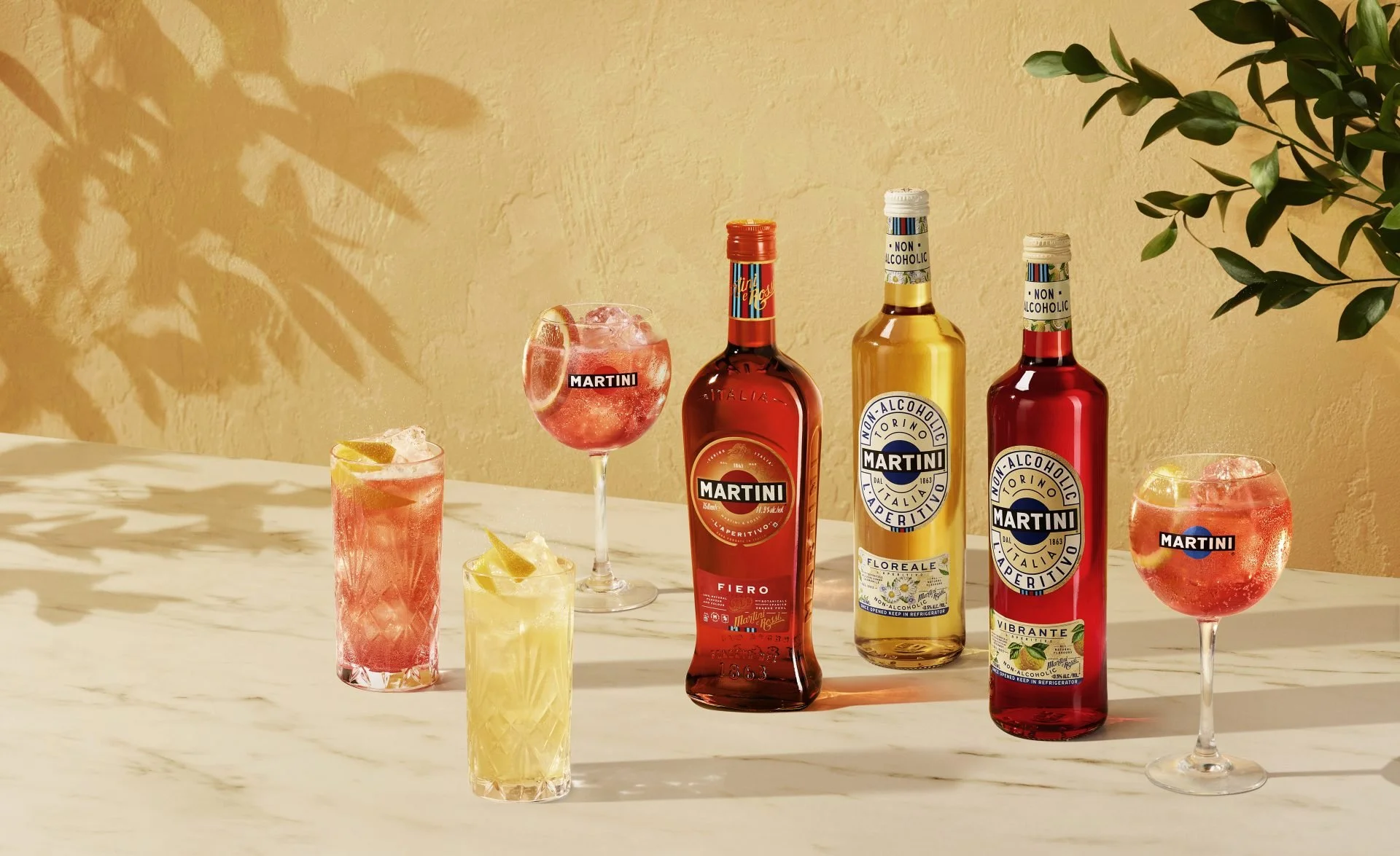 Various bottles of Martini non-alcoholic aperitif and colorful cocktails with lemon and orange slices on a marble table against a textured yellow wall, with shadows of leaves cast on the wall.