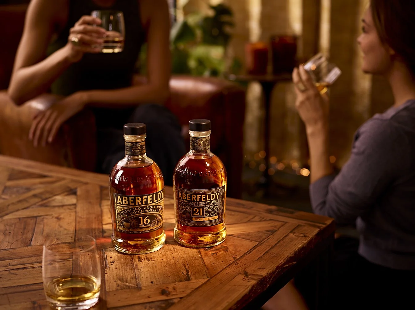 Two bottles of Aberfeldy whisky on a wooden table with a glass of whisky in the foreground. Two women are sitting in the background, holding glasses of whisky, engaged in conversation in a cozy indoor setting.