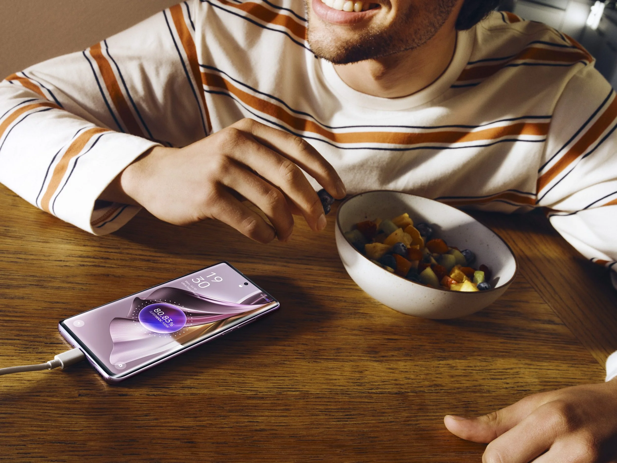 A person with a beard, wearing a striped shirt, is sitting at a wooden table. They are smiling while holding a spoon over a bowl of fruit salad. A smartphone is placed on the table, connected to a charging cable.