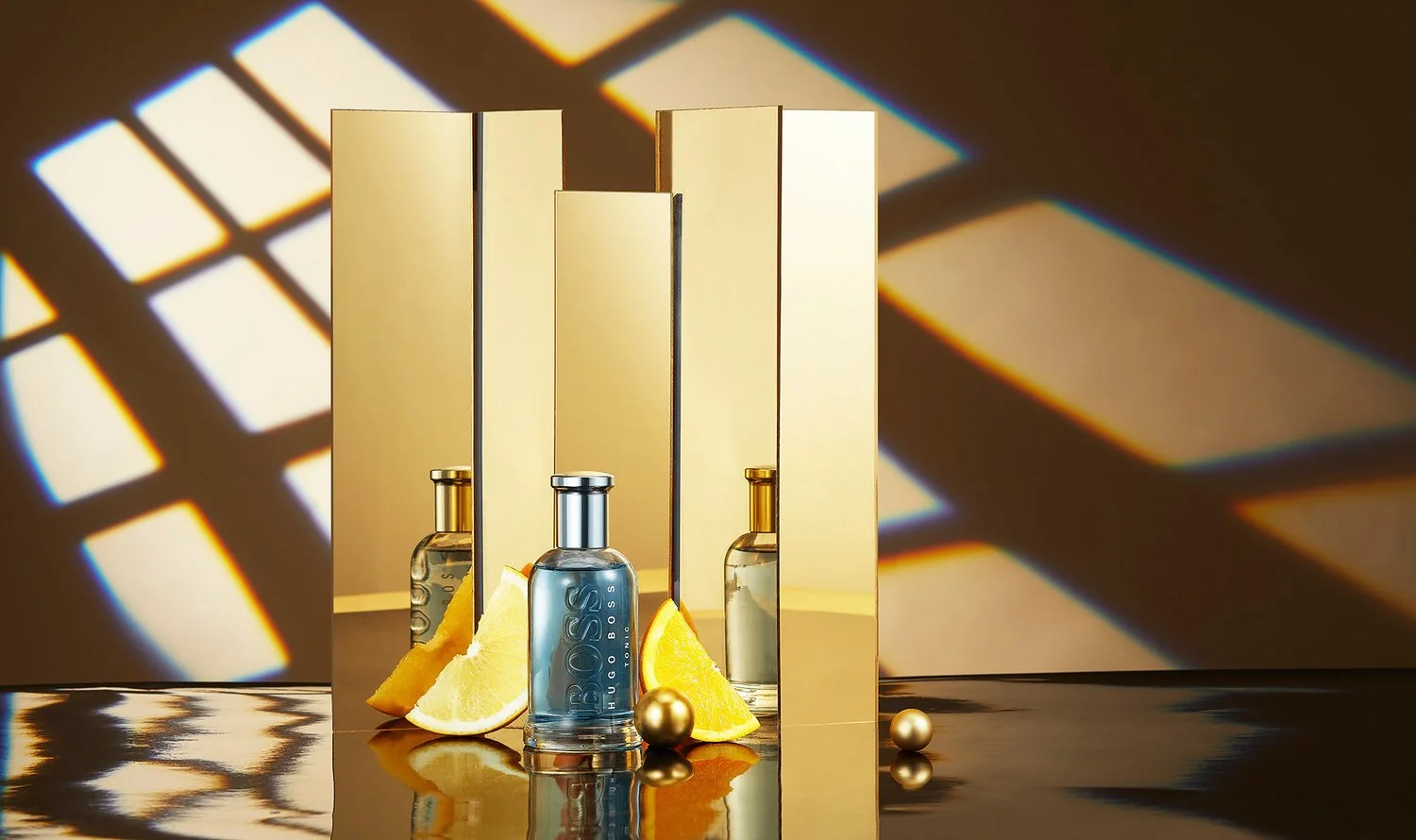 A perfume bottle with the label 'BOSS', slices of lemon, and golden spheres are displayed in front of a mirror with a geometric shadow pattern.