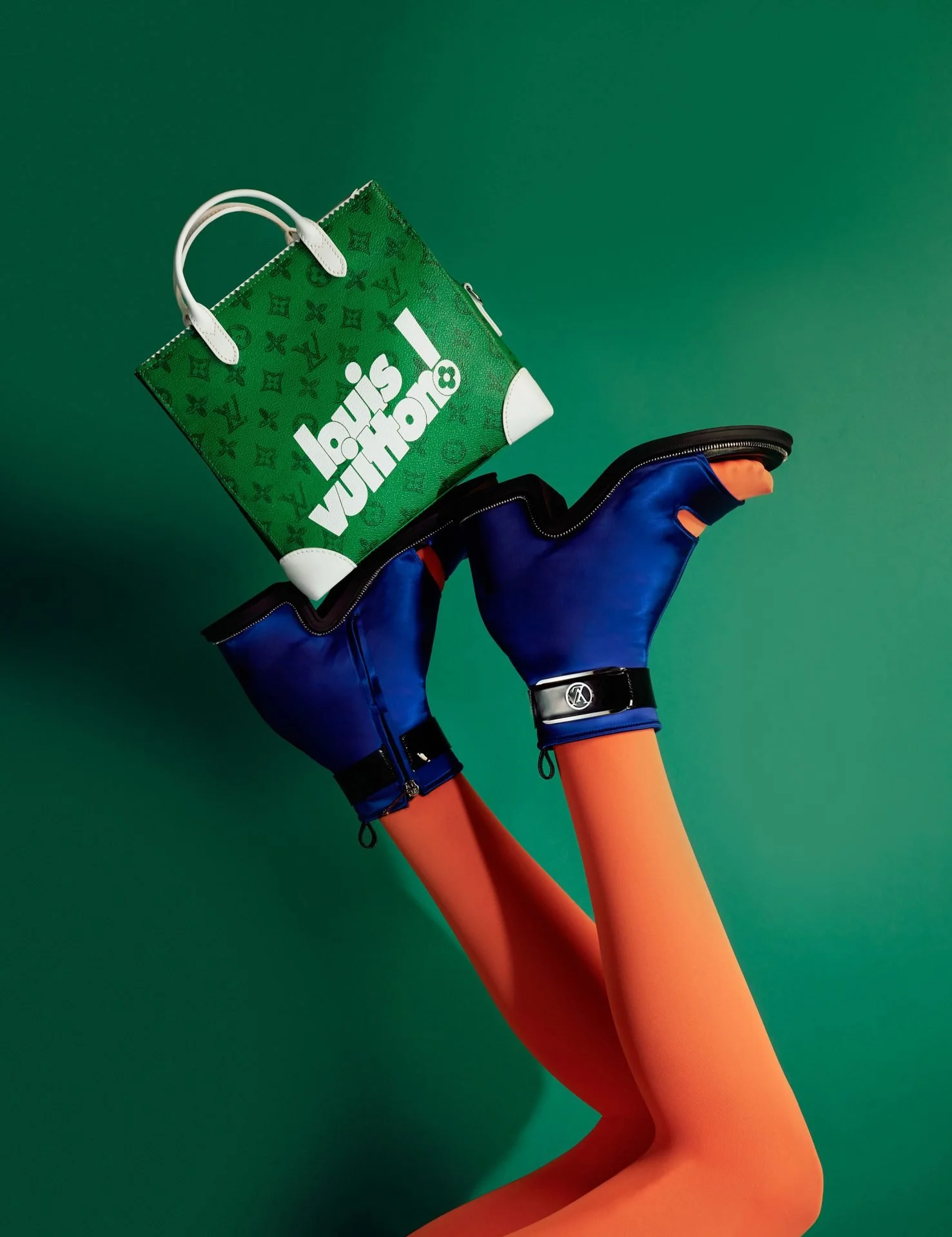 Fashion mannequin legs wearing blue high-heeled shoes with orange stockings, holding a Louis Vuitton shopping bag against a green background.