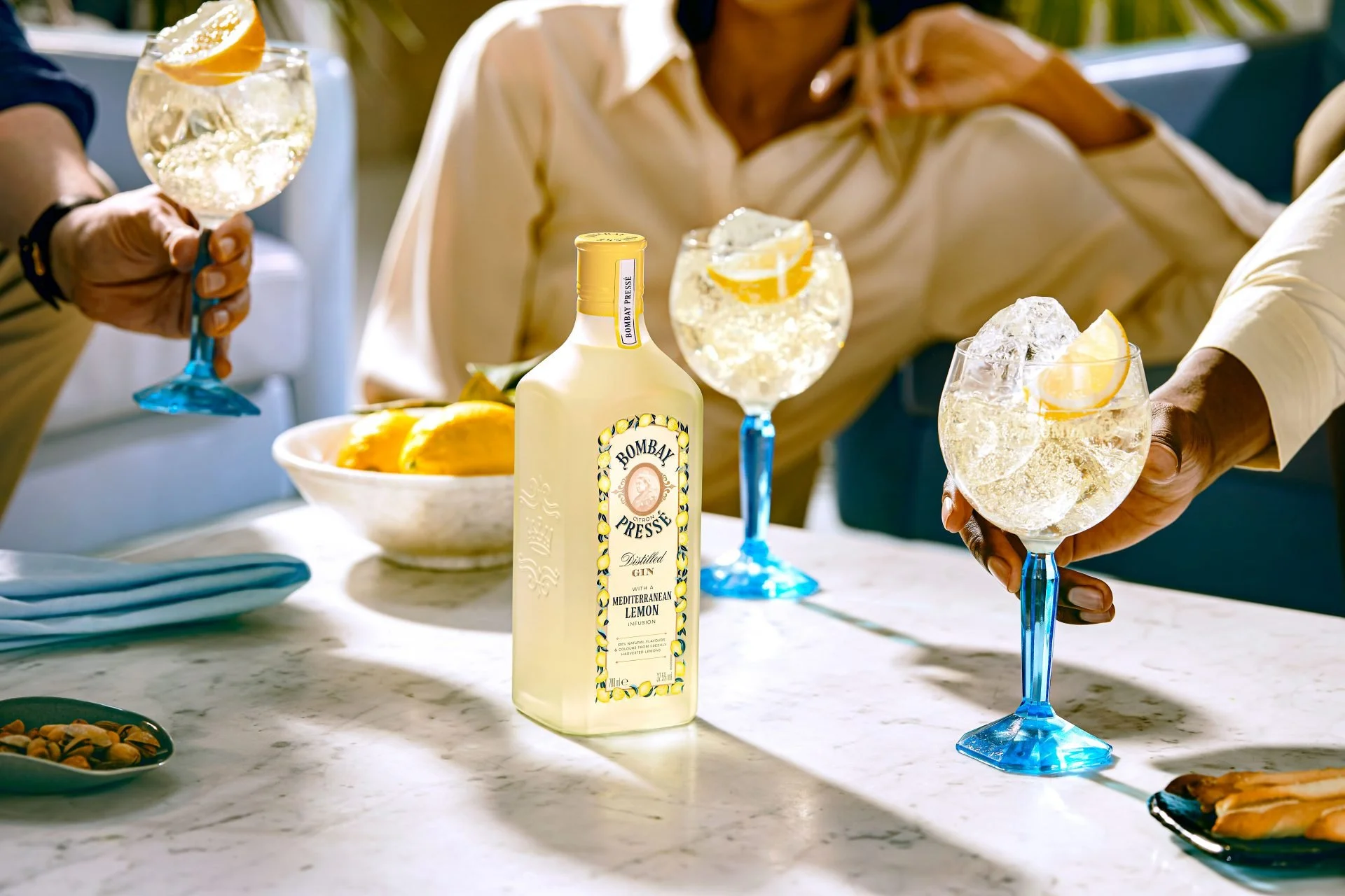 People clinking glasses of gin and lemonade at a social gathering, with a bottle of Bombay Pressé Mediterranean Lemon gin and a bowl of lemons on a white marble table.