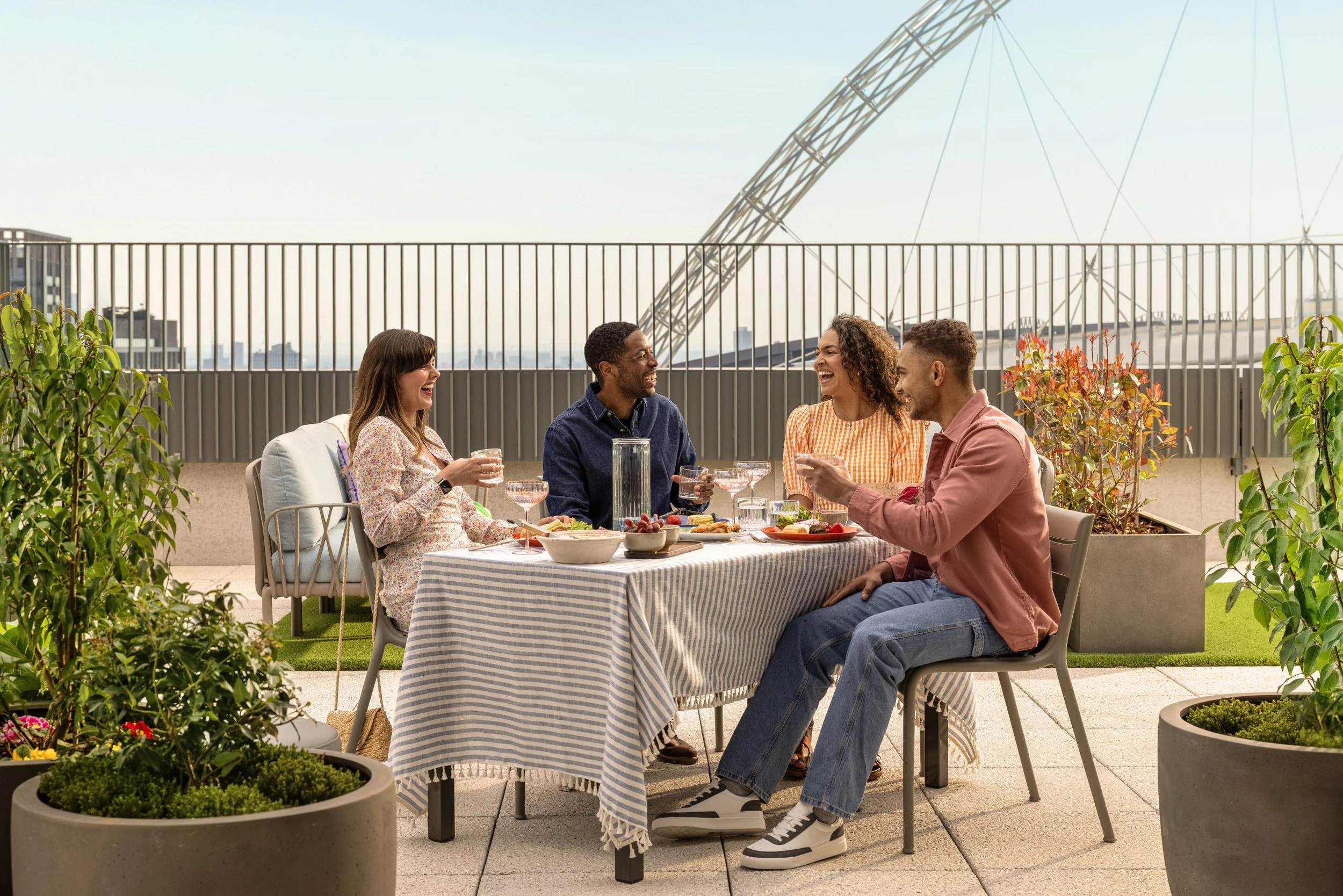 Four friends sitting at a dining table on a rooftop terrace, enjoying a meal and drinks, with city buildings and a ferris wheel in the background.