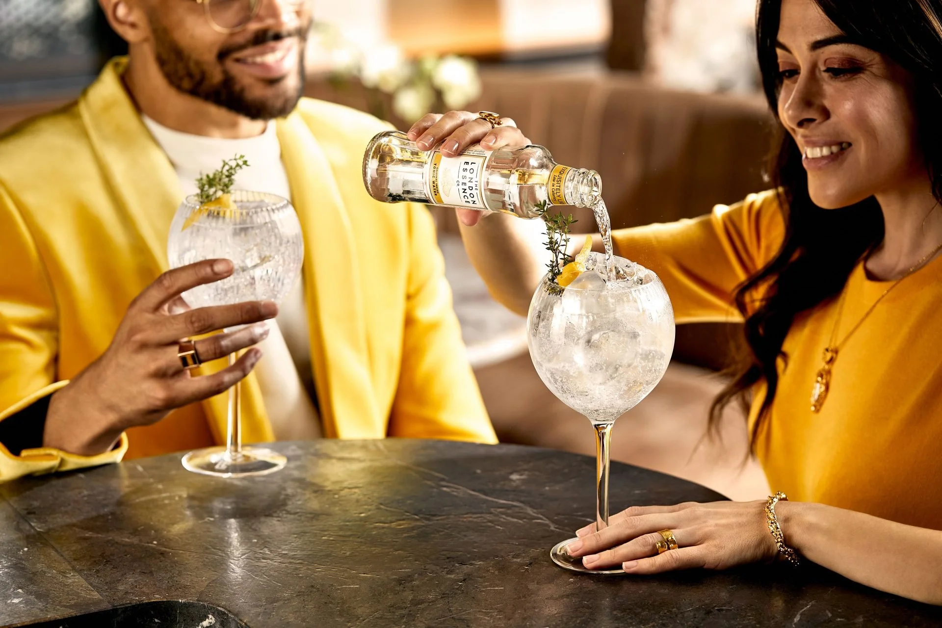 A man and woman sitting at a bar or restaurant, smiling and dressed in yellow, sharing a moment as the man pours a clear beverage with herbs and lemon into a large glass with ice, garnished with herbs and lemon slices.