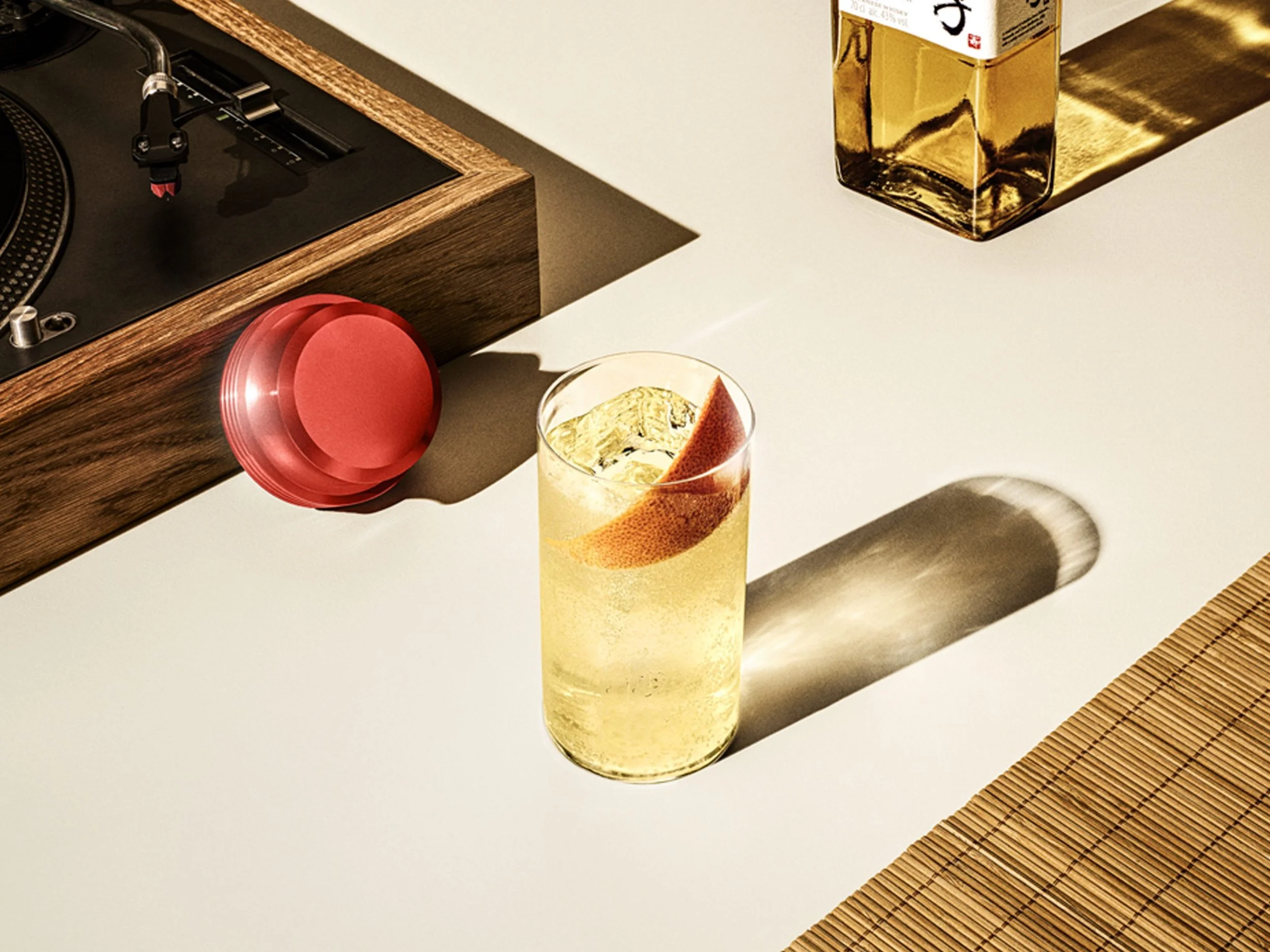 A glass of a yellow beverage with ice and a slice of grapefruit on the rim, placed on a white surface. In the background, a golden paper bag and part of a wooden speaker with a turntable are visible.