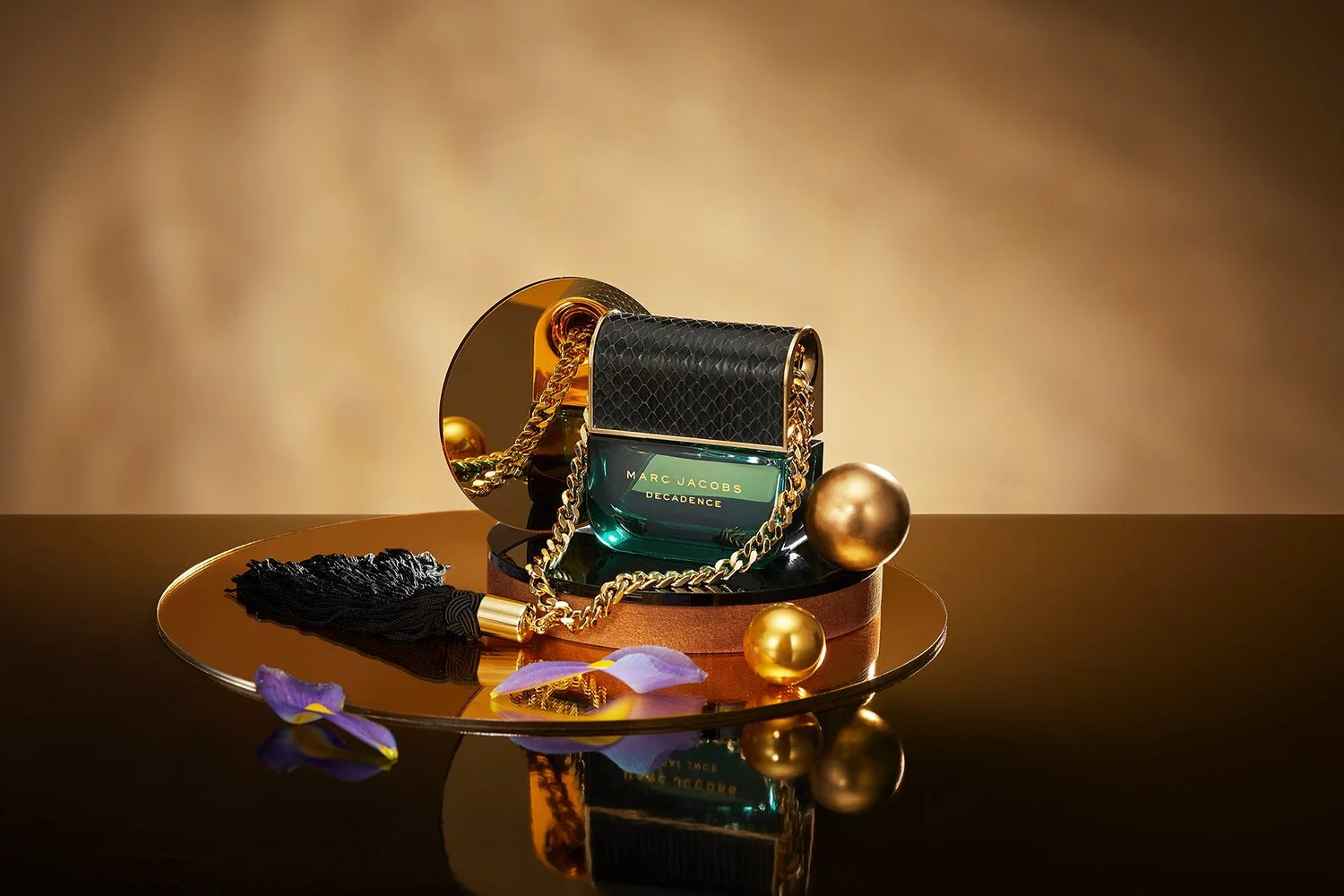 A Marc Jacobs Decadence perfume bottle surrounded by gold decorative spheres, a black tassel, and purple flower petals on a reflective surface with a golden background.