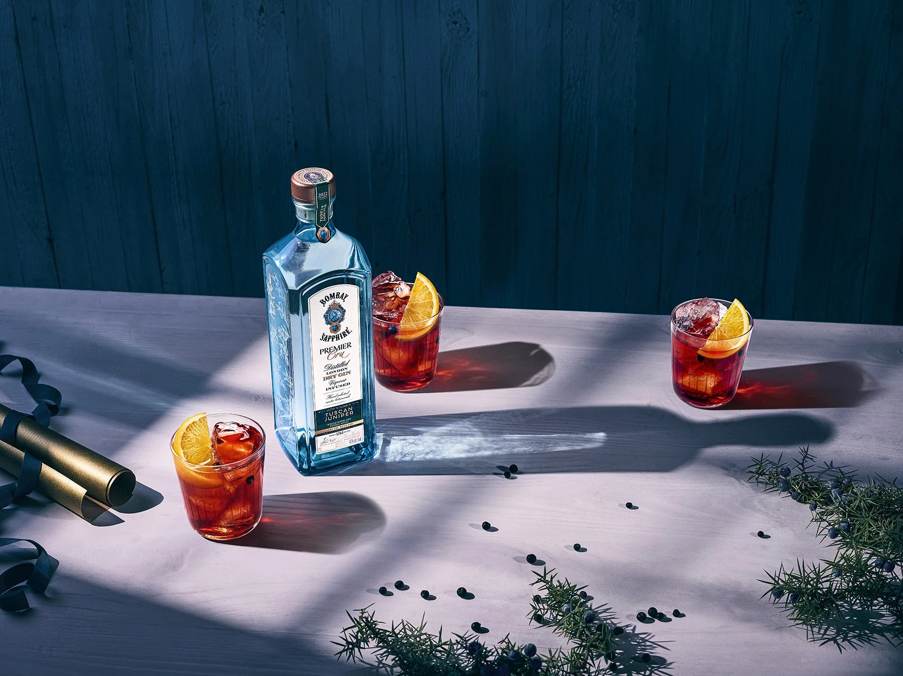 A bottle of Bombay Sapphire gin and three glasses of red drink with ice and lemon slices on a white tablecloth, outdoor setting with shadows and scattered coffee beans and greenery.