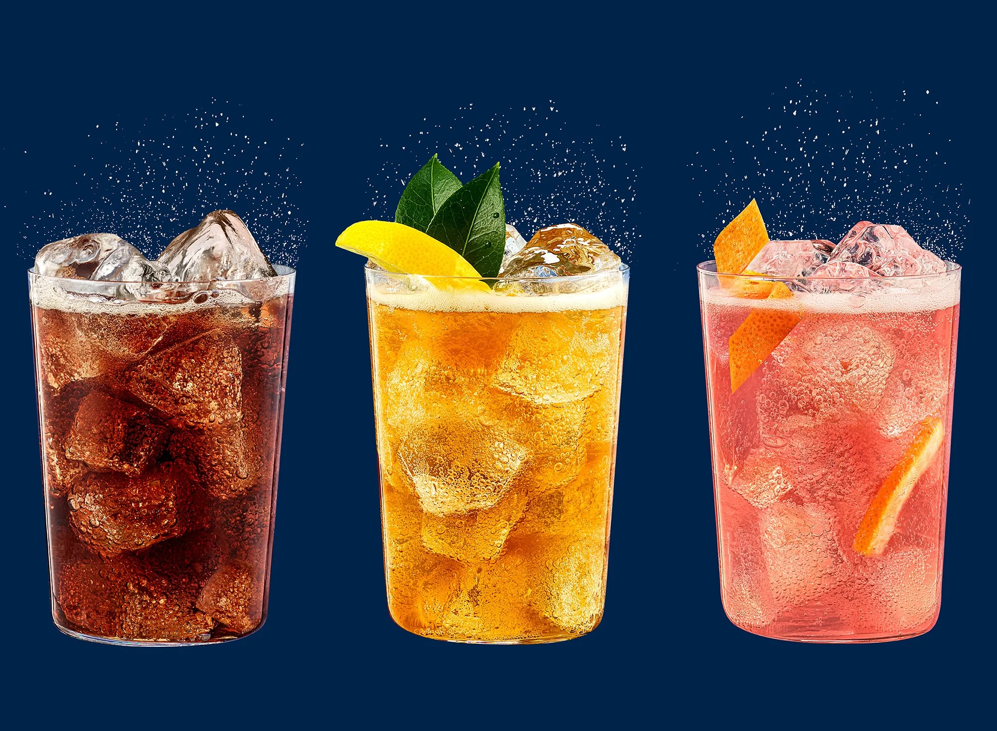 Three tall glasses filled with colorful soda drinks, each with ice and garnished with lemon, lime, and orange slices, and stirred with sprinkled sugar or salt on top, against a dark blue background.