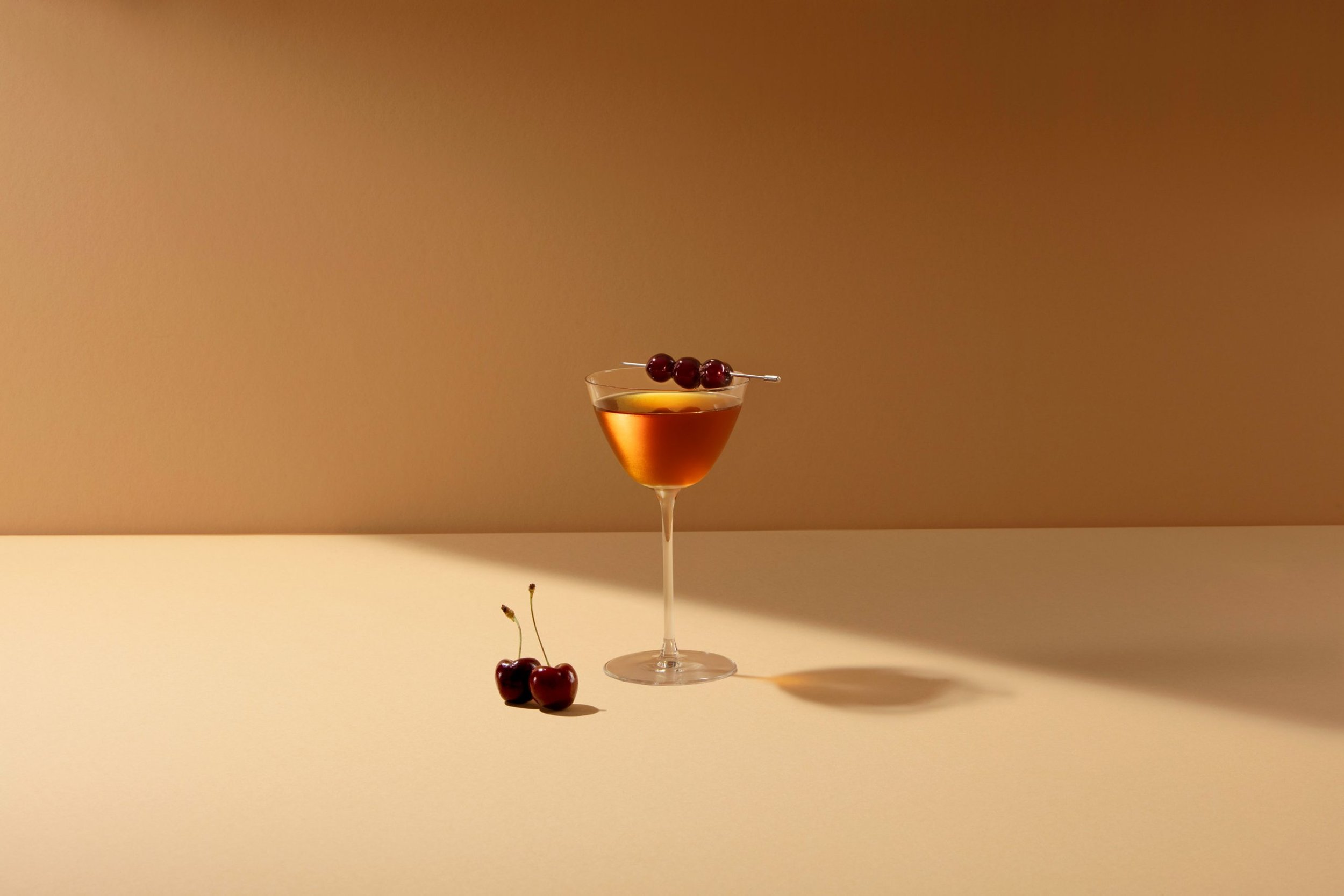 A cocktail glass with an orange-colored drink garnished with three cherries on a skewer. There are two cherries on the surface below the glass, and a single cherry with a stem is on the far left.