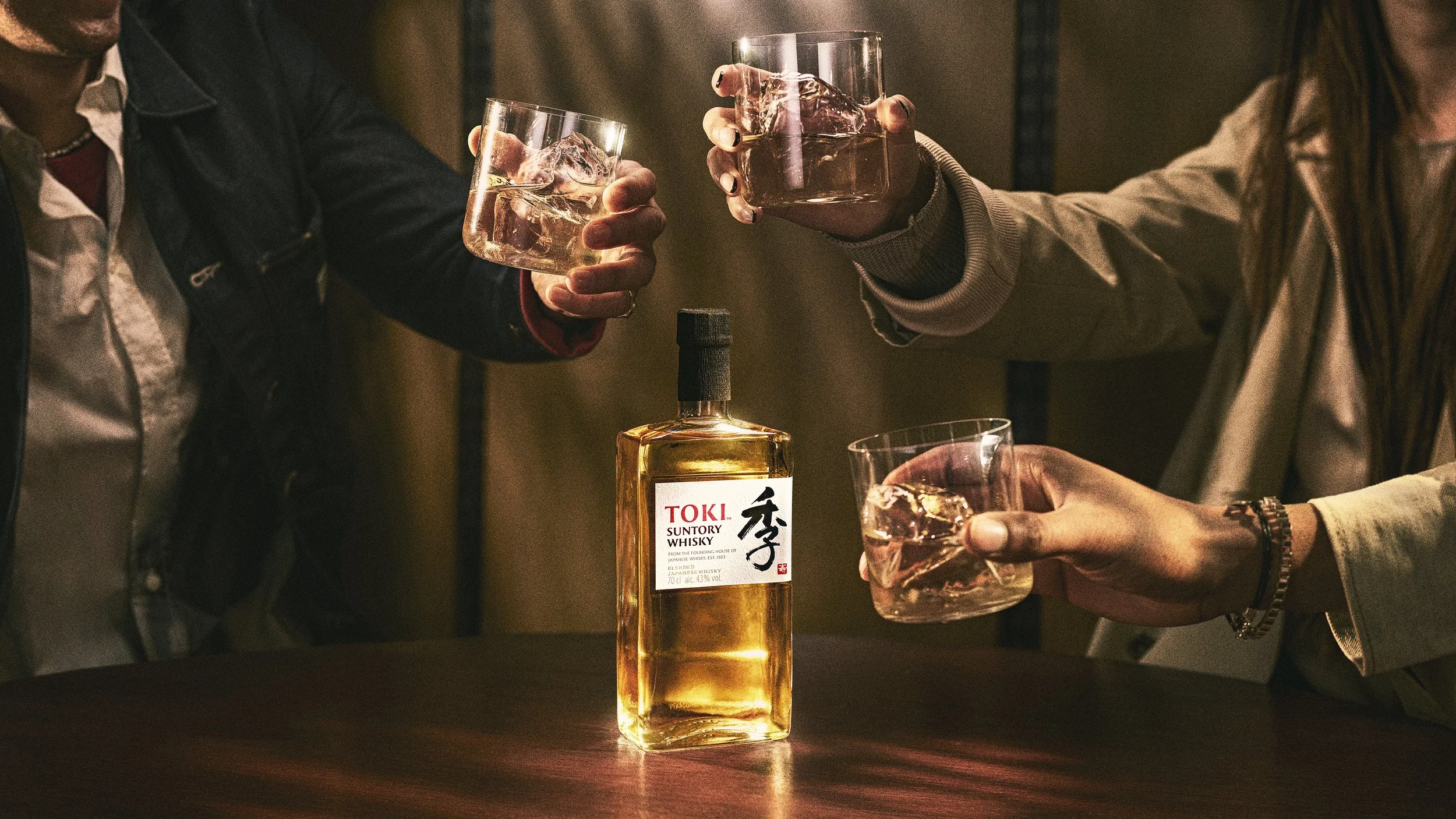 Three people toasting with glasses of whiskey with ice, with a bottle of Toki Suntory Whisky on a wooden table.
