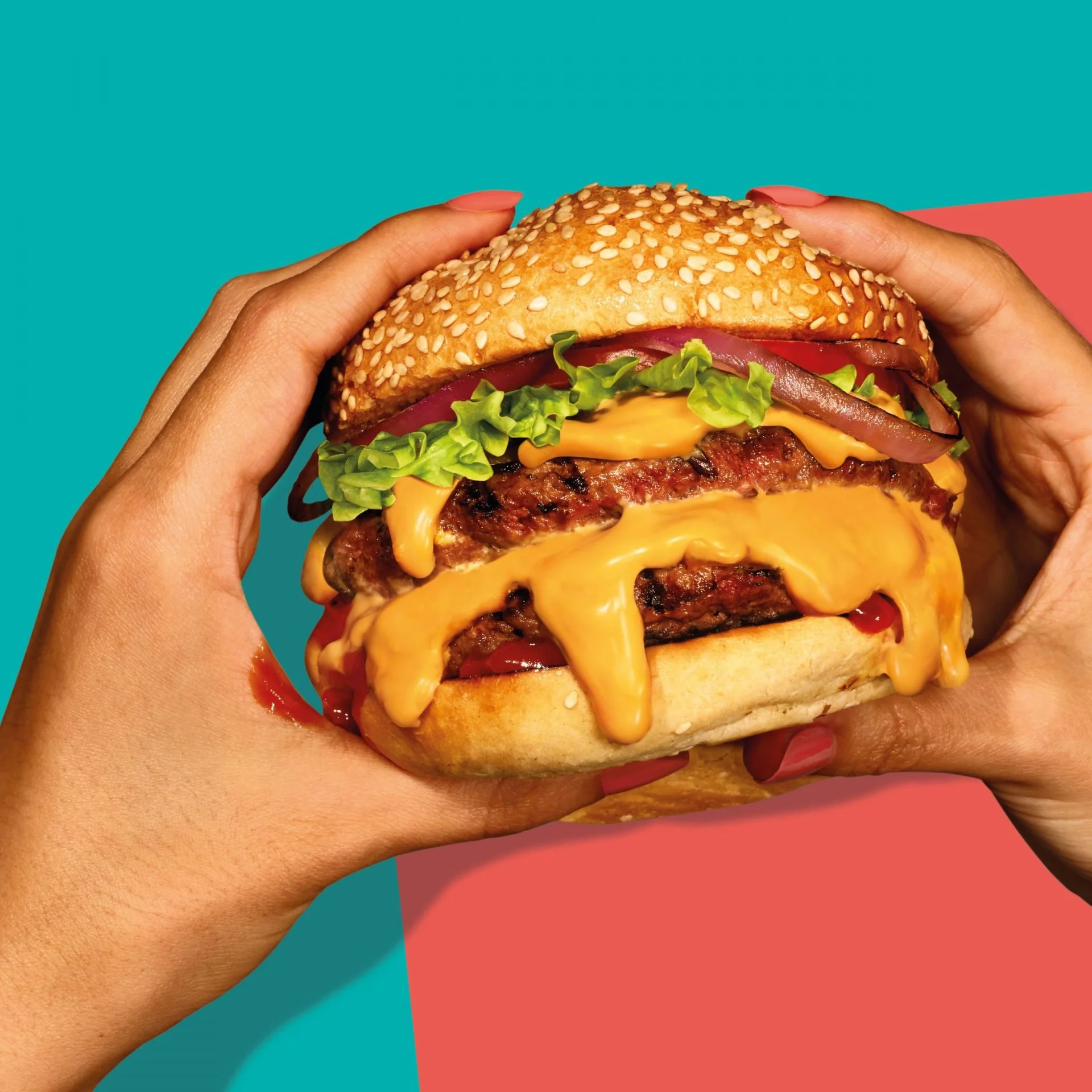 Close-up of a person holding a cheeseburger with lettuce, tomato, bacon, cheddar cheese, and a sesame seed bun against a split teal and pink background.