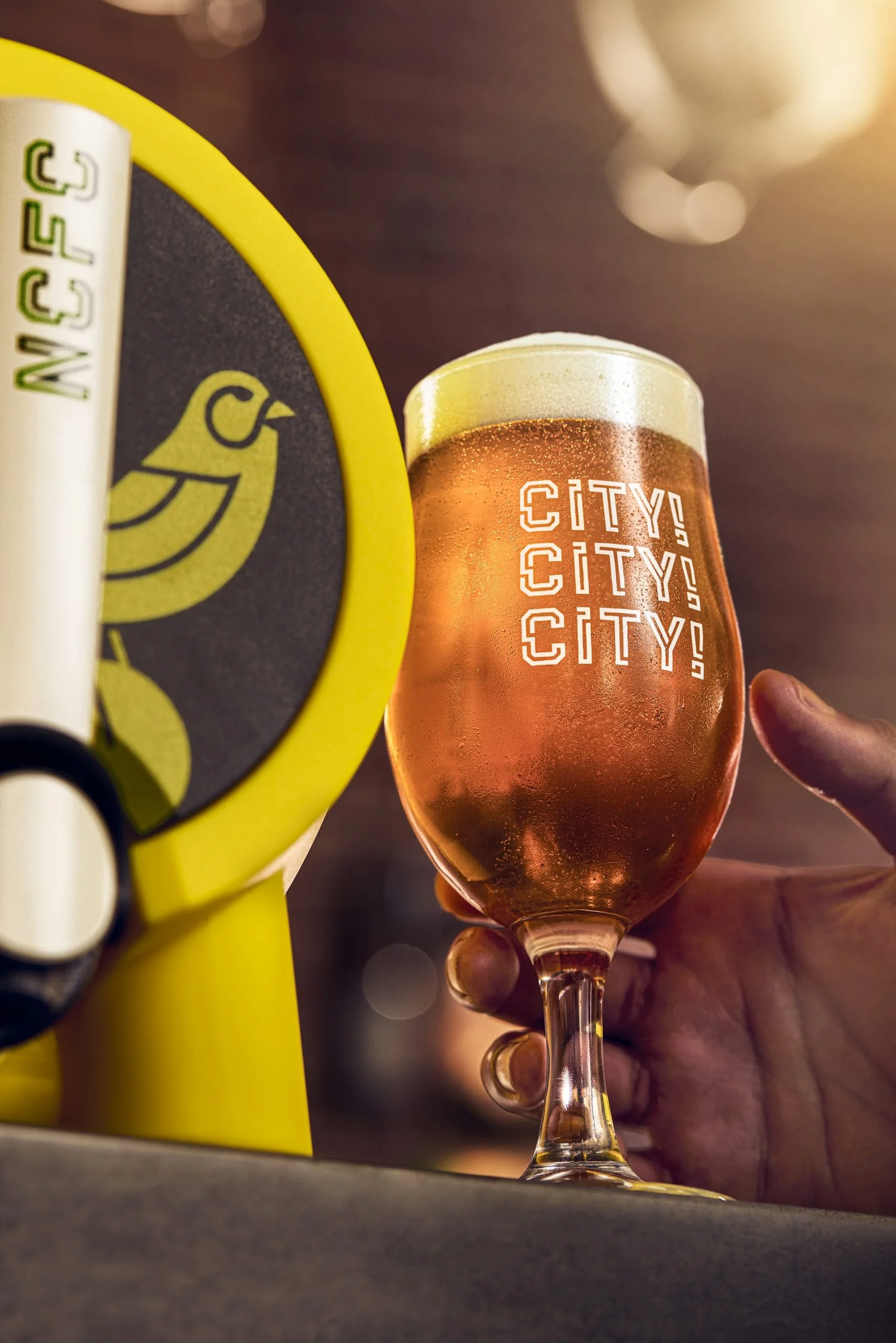 Close-up of a glass of pinkish beer with the word 'CITY!' printed in white, held by a person's hand with a ring on their finger. Part of a yellow and black object with a bird logo is visible on the left side, and the background is blurred with warm l