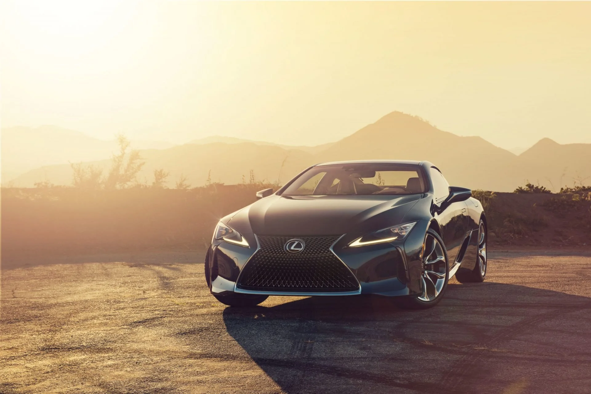 A black Lexus convertible sports car parked on a dirt road during sunset with mountains and sparse vegetation in the background.
