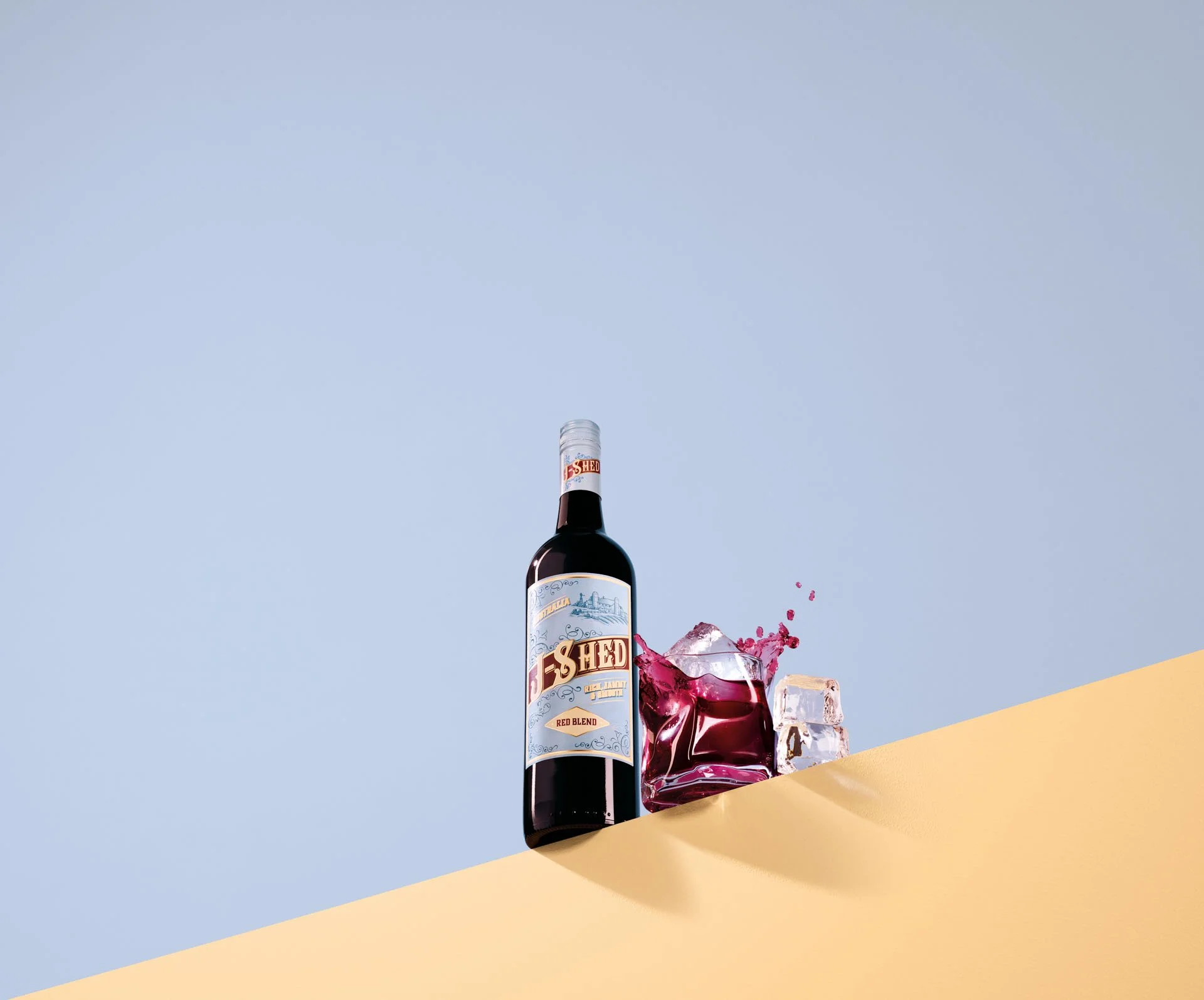 A bottle of red wine labeled 'Shed' standing on a yellow surface next to a glass filled with red wine, ice cubes, and some splashes of red wine with ice cubes in mid-air against a light blue background.