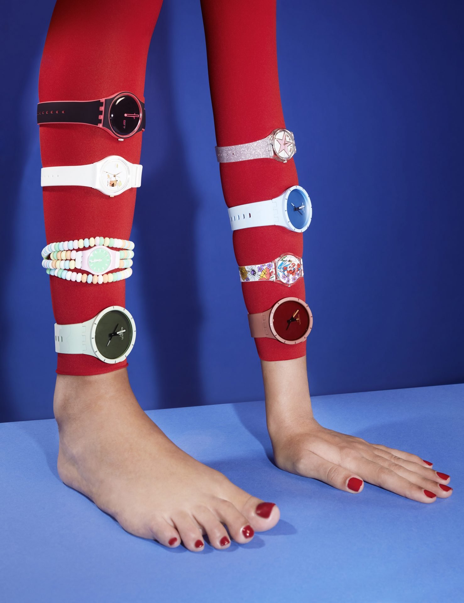 Person in a yoga pose with both arms and hands flat on the blue surface, wearing colorful watches on their forearms, red nail polish, and a red outfit / sleeves against a blue background.