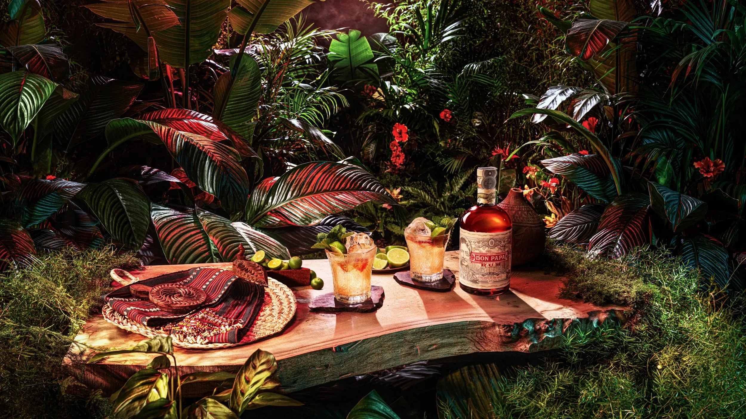 A tropical setting with lush green leaves and plants, with a table holding a bottle of Don Papa rum, cocktails with lime slices, and decorative items including woven textiles and pottery.